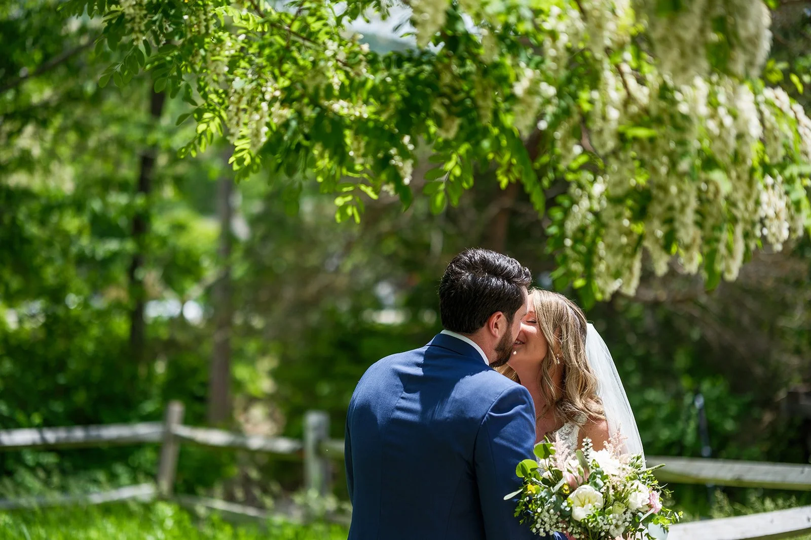 A bride and groom share a kiss outdoors in a lush green setting, with the bride holding a bouquet of flowers and sunlight filtering through the trees.