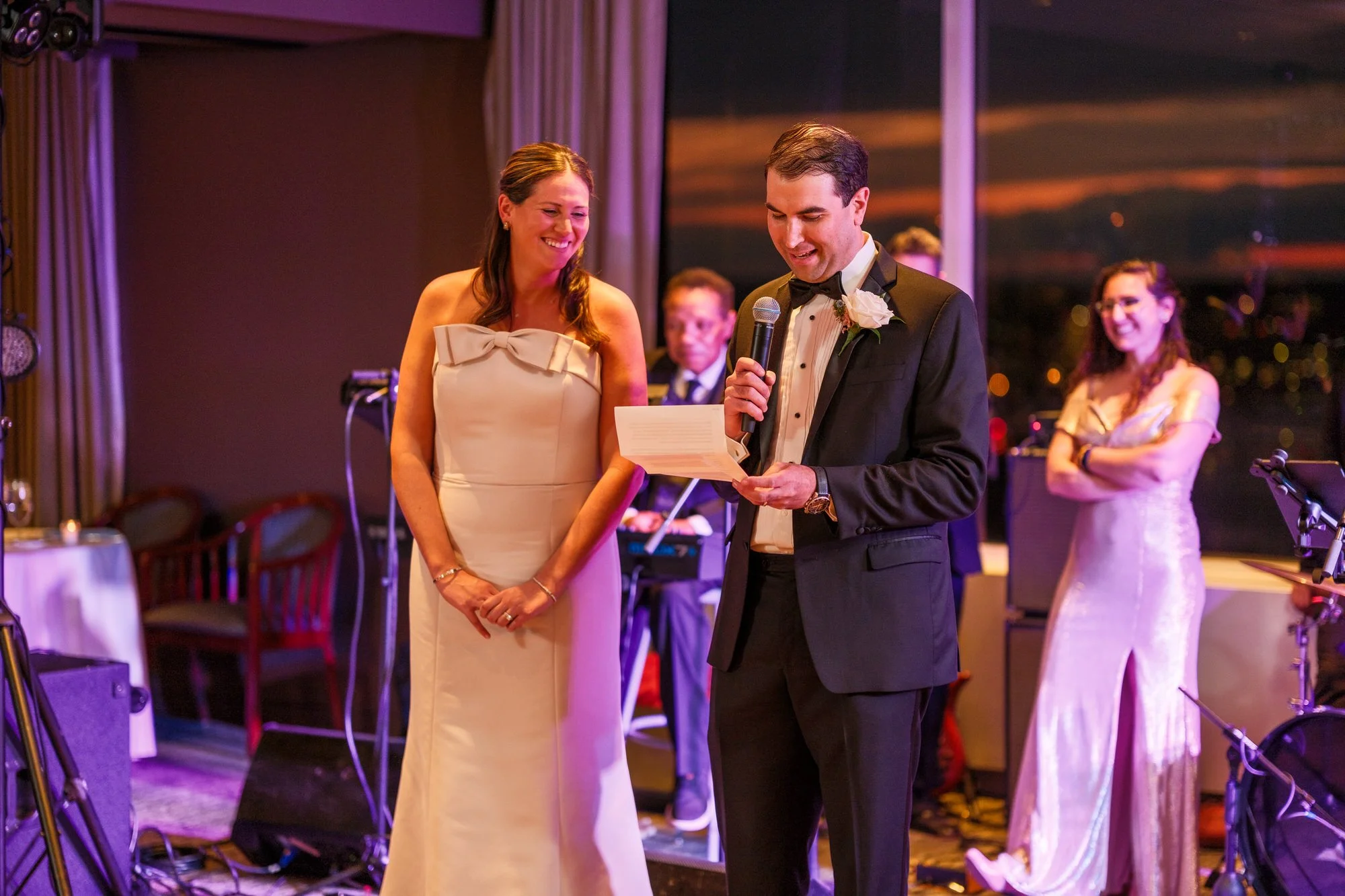 Best man giving a toast while holding notes and microphone next to the bride at the UMass Club Boston wedding reception