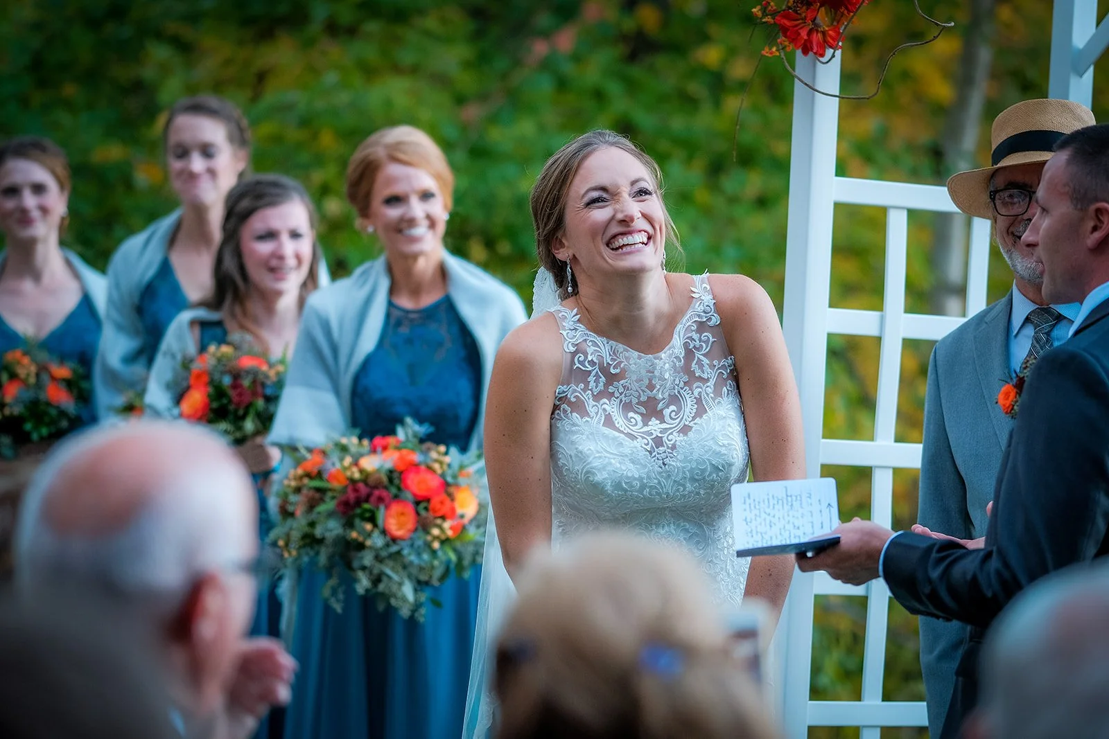 Bride smiling during her wedding vow exchange outdoors with bridesmaids and guests in background.