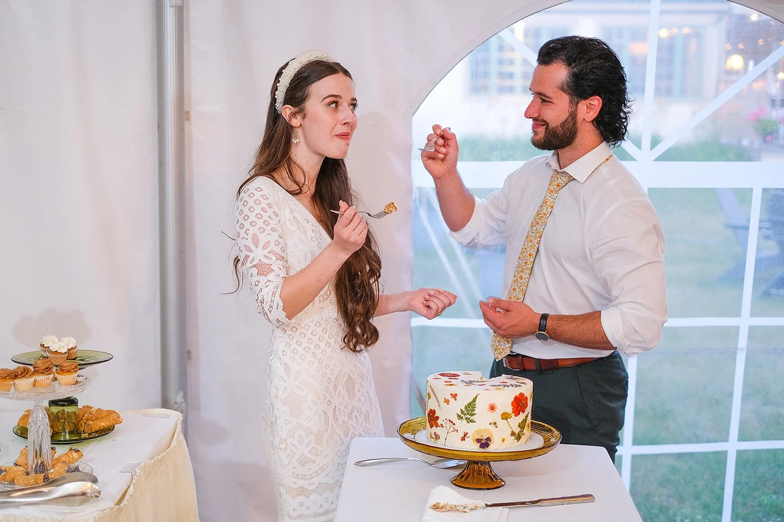 A woman in a white lace dress and a headband is holding a fork with cake, standing next to a man in a white shirt and patterned tie, who is feeding her with a fork. There is a decorated cake on a table between them, and a window with city buildings o