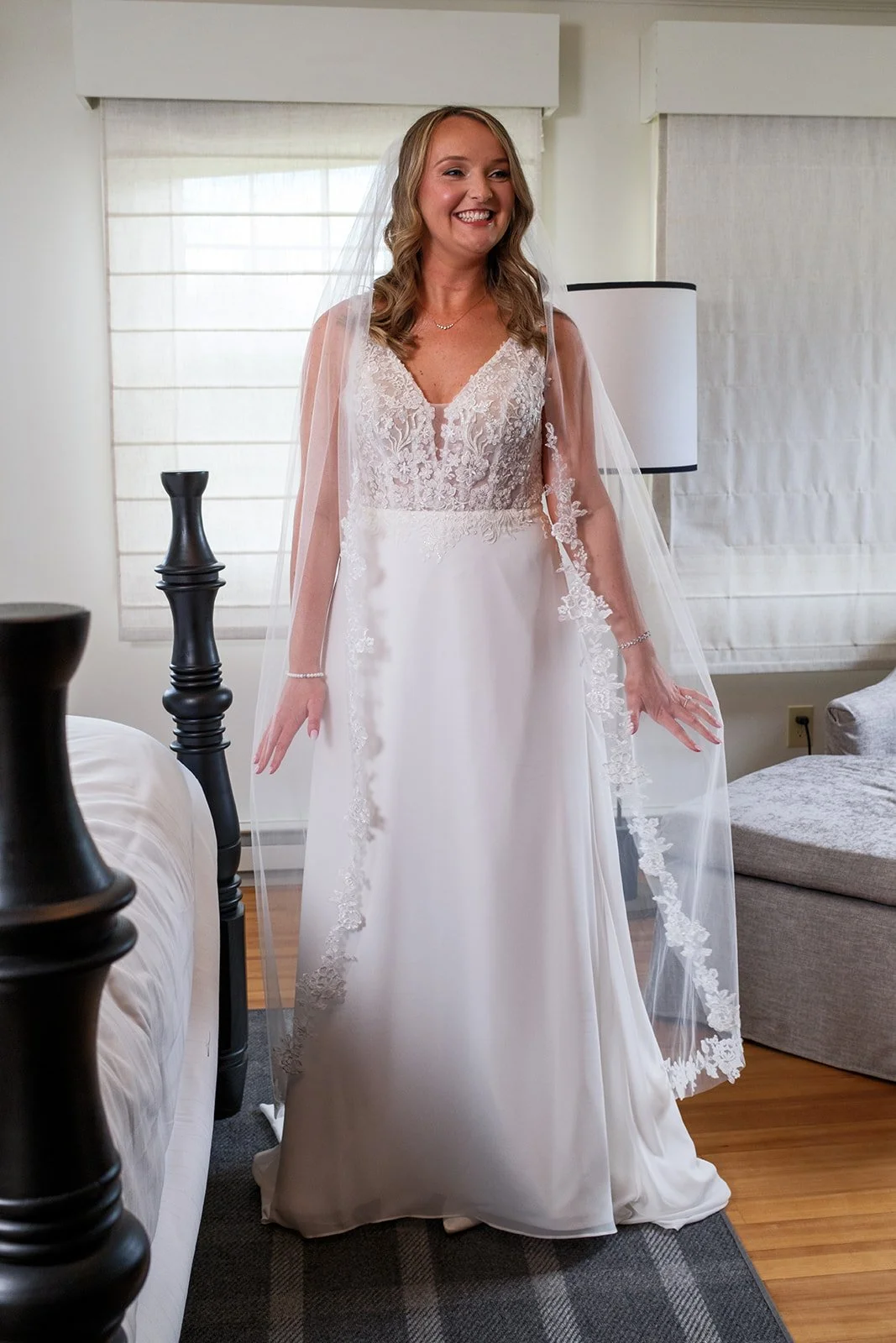 A smiling woman in a wedding dress with lace details on the bodice and a flowing skirt, standing indoors near windows and furniture.