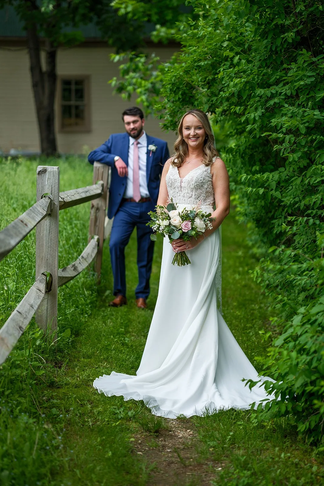 A bride in a white wedding dress holding a bouquet of flowers, standing on a grassy pathway next to a wooden fence, with a groom in a blue suit and tie leaning on the fence behind her, surrounded by greenery.