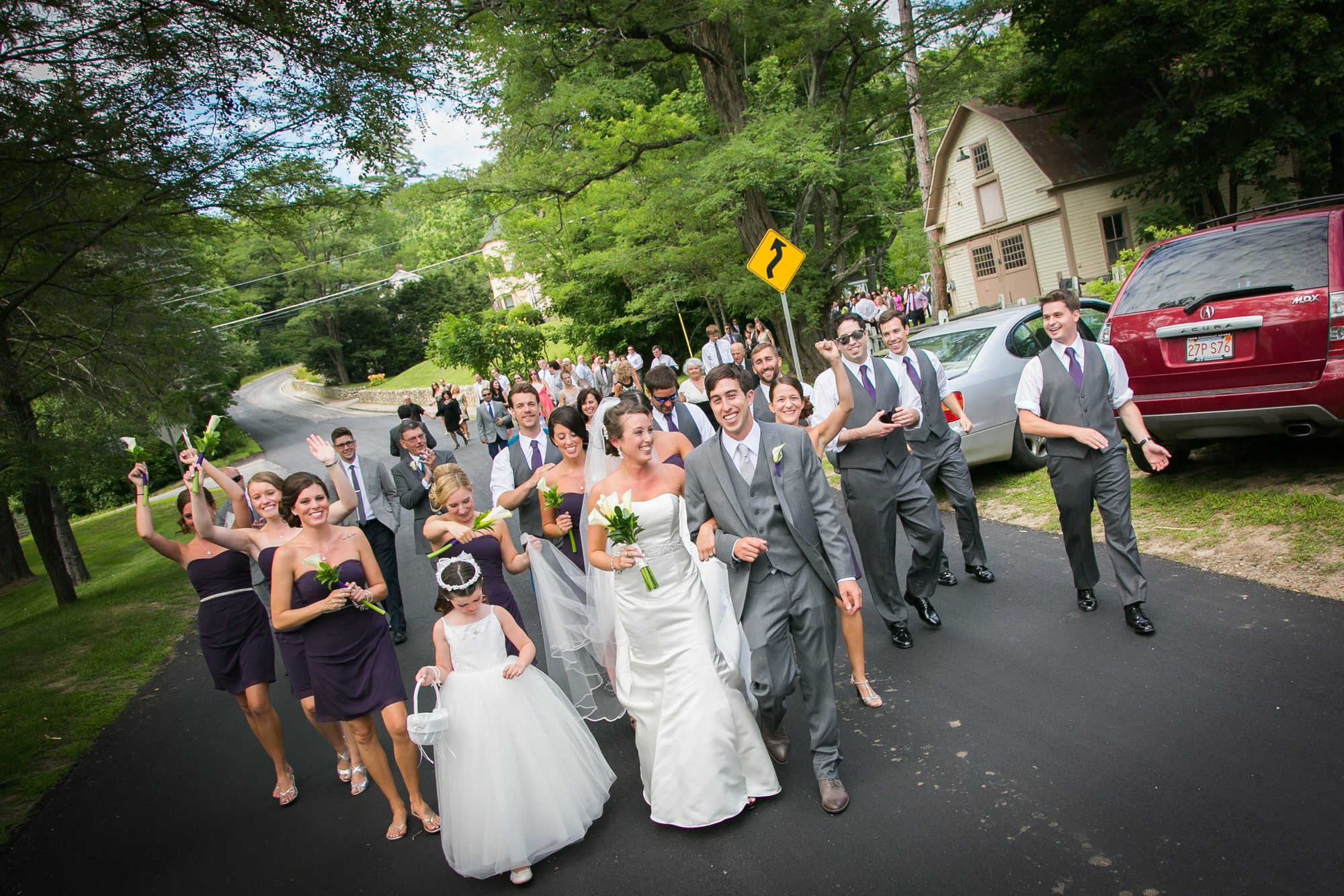 Bride and groom leading a wedding procession of bridesmaids and groomsmen walking on a suburban street, smiling and waving, with many guests in the background.
