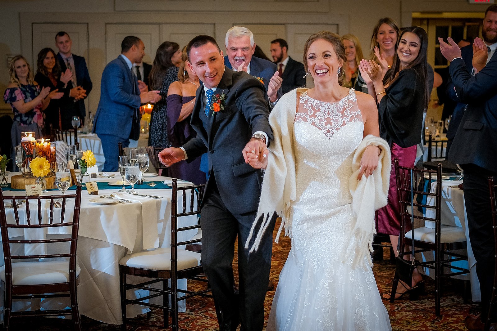 Bride and groom holding hands and walking through a reception hall, smiling, surrounded by guests clapping and celebrating, decorated with flowers and candles on tables.
