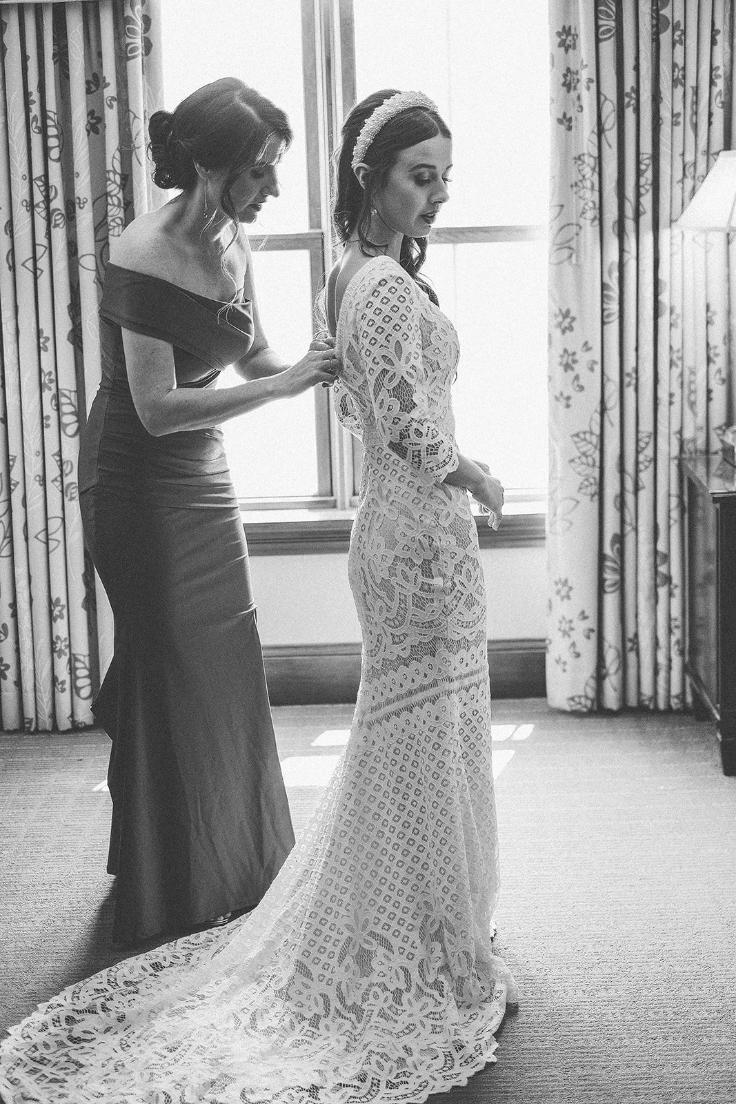 A woman in a lace wedding dress is being assisted with her dress by another woman in an off-shoulder gown, inside a room with floral curtains and a window.