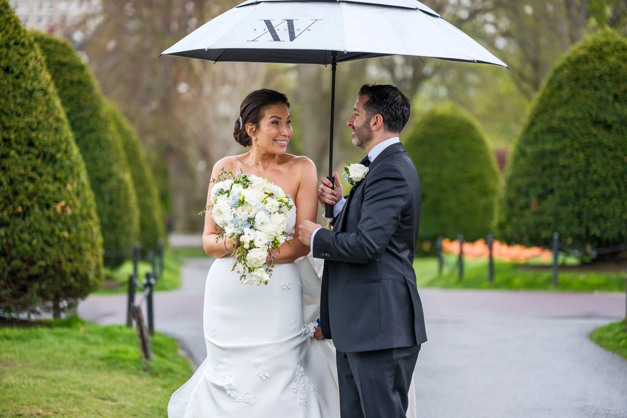 Bride and groom laughing under an umbrella in the Boston Public Garden on a rainy wedding day