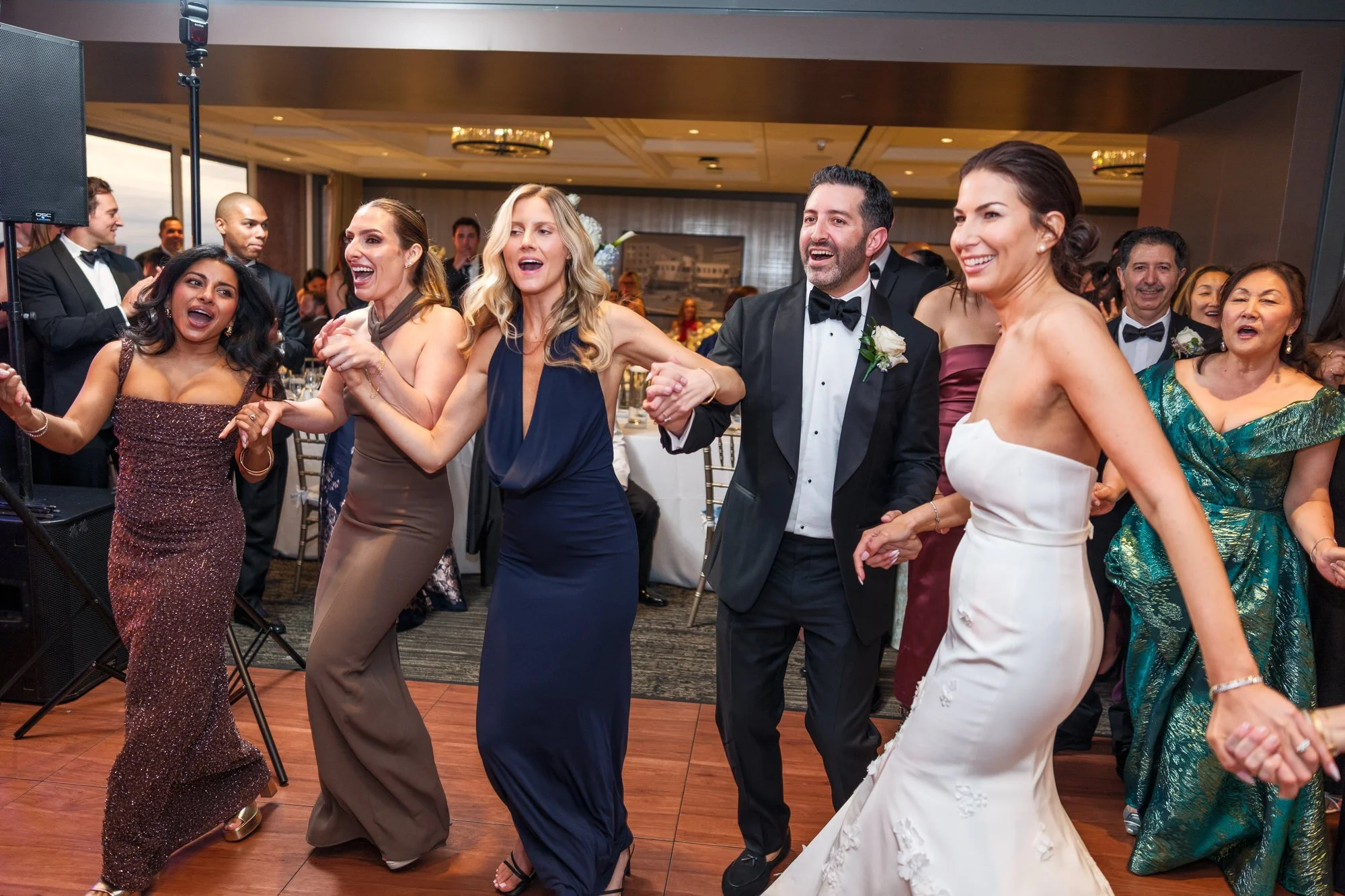 Bride, groom, and guests dancing in a joyful line during the hora at the UMass Club Boston Jewish wedding reception