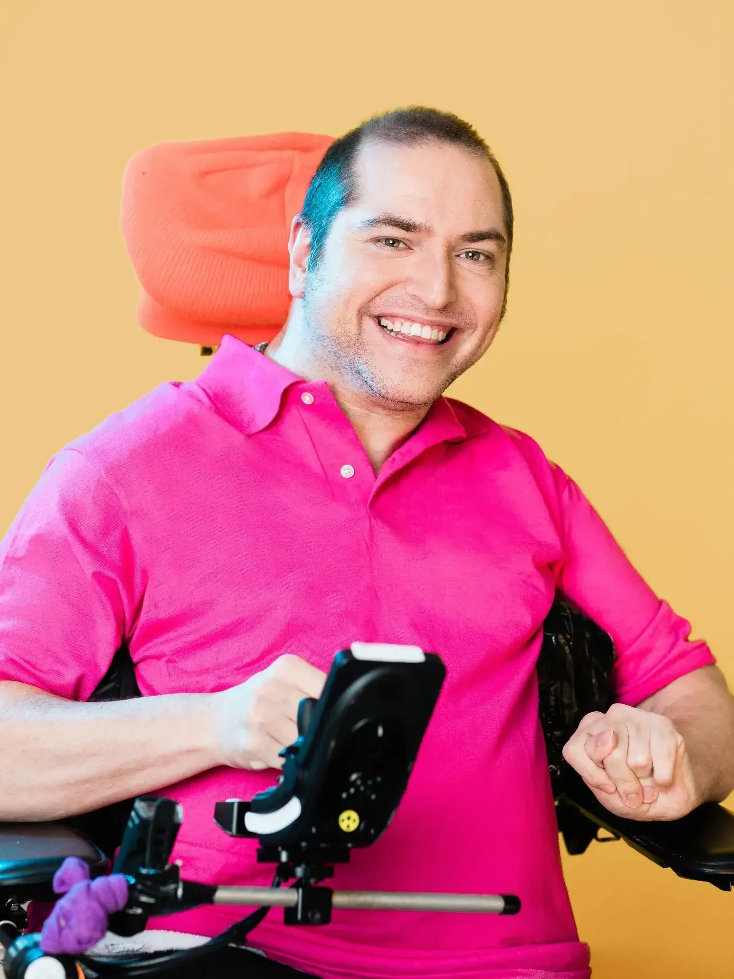Andrew Gurza is wearing a bright pink polo shirt and he is sitting in a black wheelchair with a headrest covered by an orange knit hat