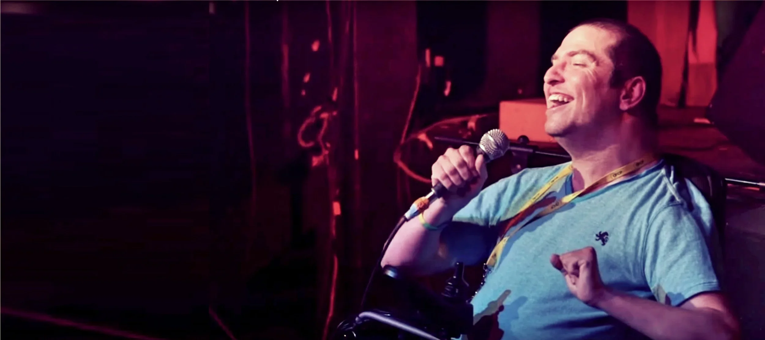 Andrew Gurza is wearing a light blue t-shirt and he is sitting in a black wheelchair with a headrest and holding a microphone while he speaks on stage