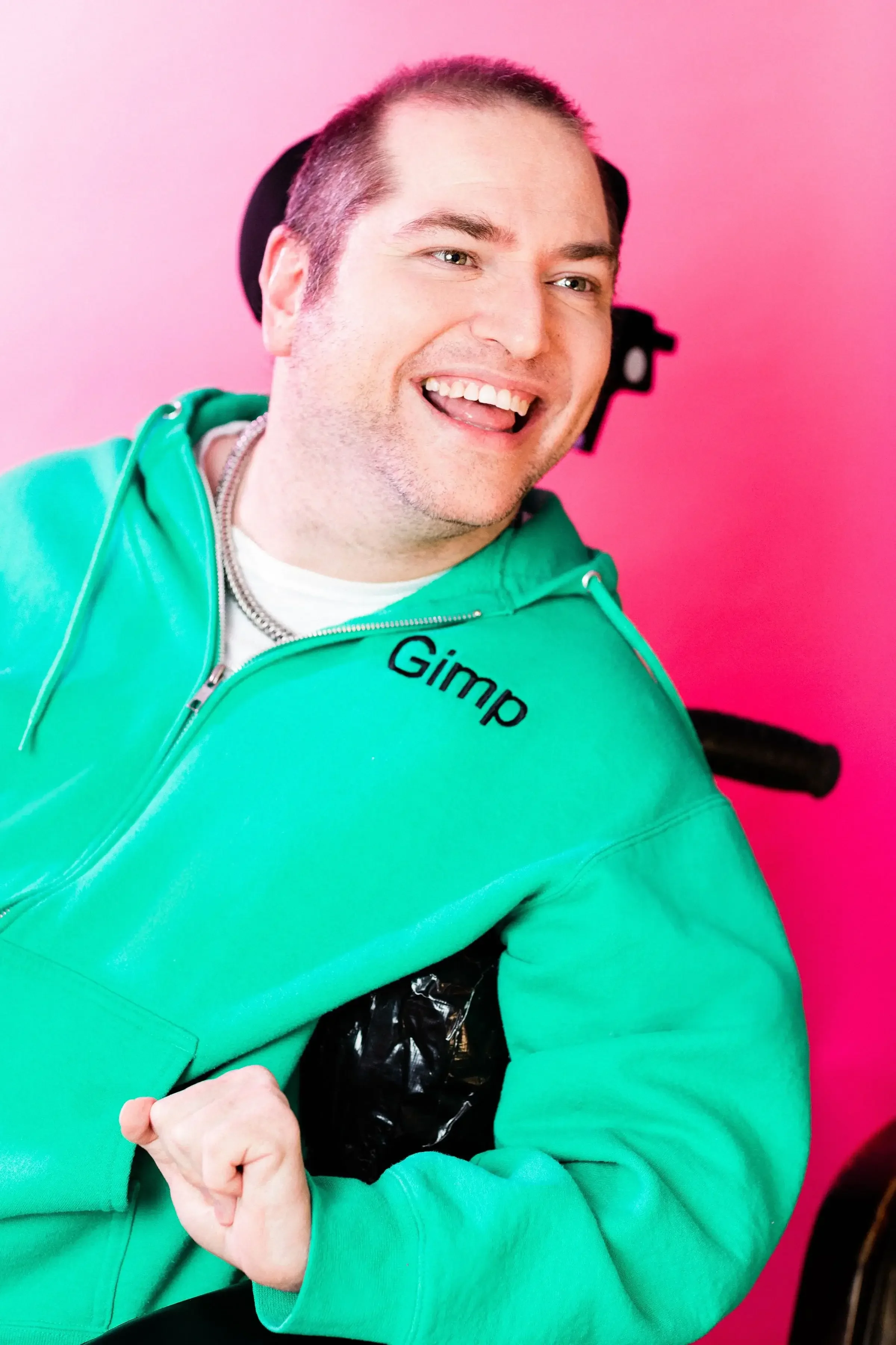 Andrew Gurza is smiling and wearing a bright green zip-up hoodie with Gimp stitched on the right side and he is sitting in a black wheelchair with a headrest
