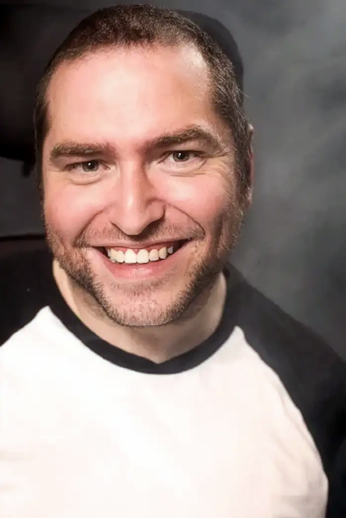 Andrew Gurza is smiling and wearing a shirt with a white body and black arms and neck.