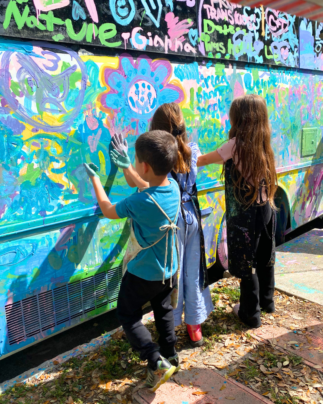 Three children with hands covered in paint leaving hand prints on the painted bus.