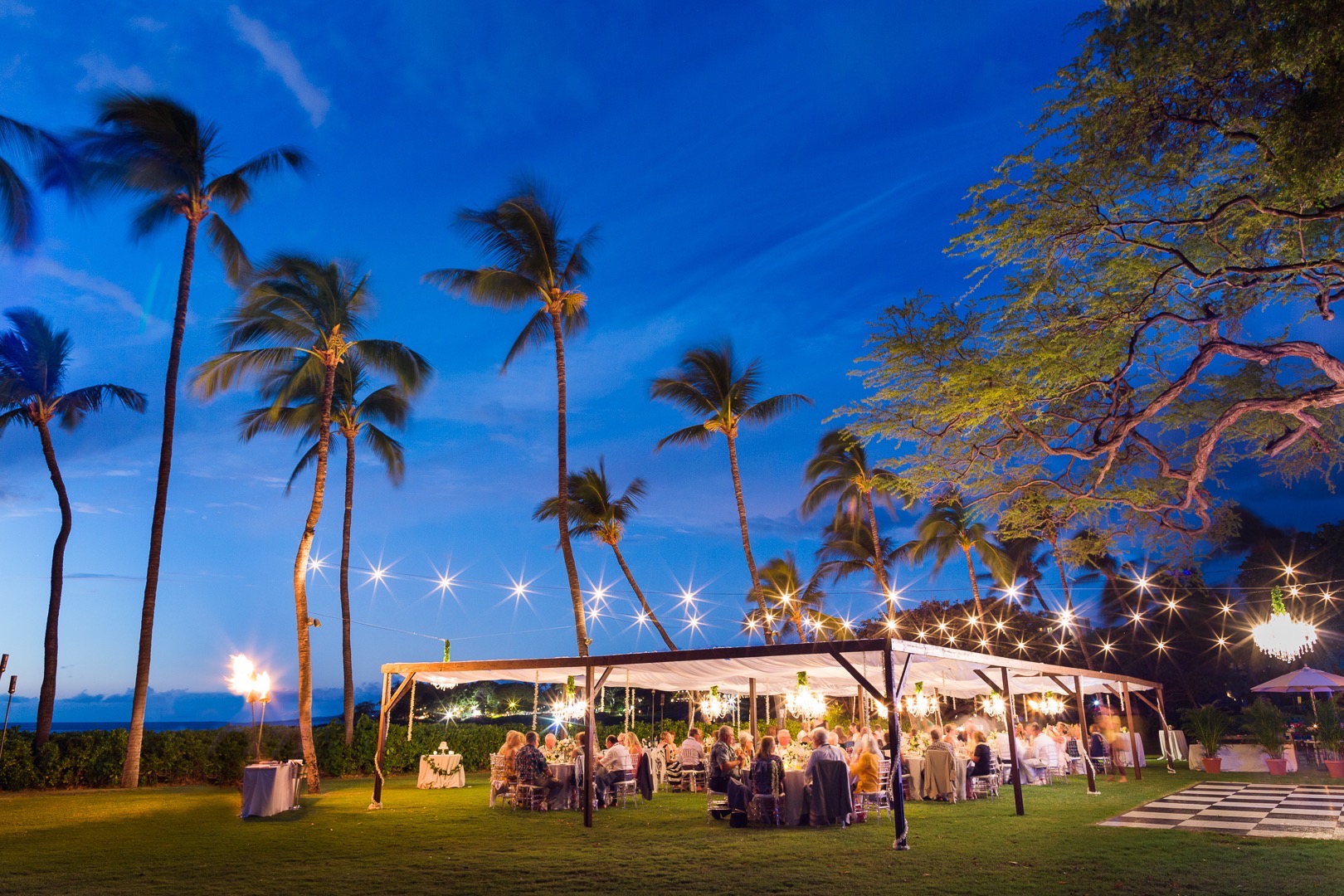 Market Lighting over our dark wood dining structure with small chandeliers and one large chandelier over the black and white dance floor for wedding at Mauna Kea Beach Hotels beautiful South point Lawn. Event design and Coordination by Bliss In Bloom | Photo Sea Light Studios .