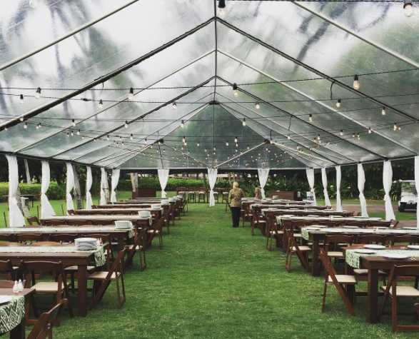 40x clear tent with thin white leg drapes, dark wood farm tables, dark wood padded folding chairs, and bistro lighting for wedding at Mauna Kea beach south point lawn. Coordinated by Bliss in Bloom