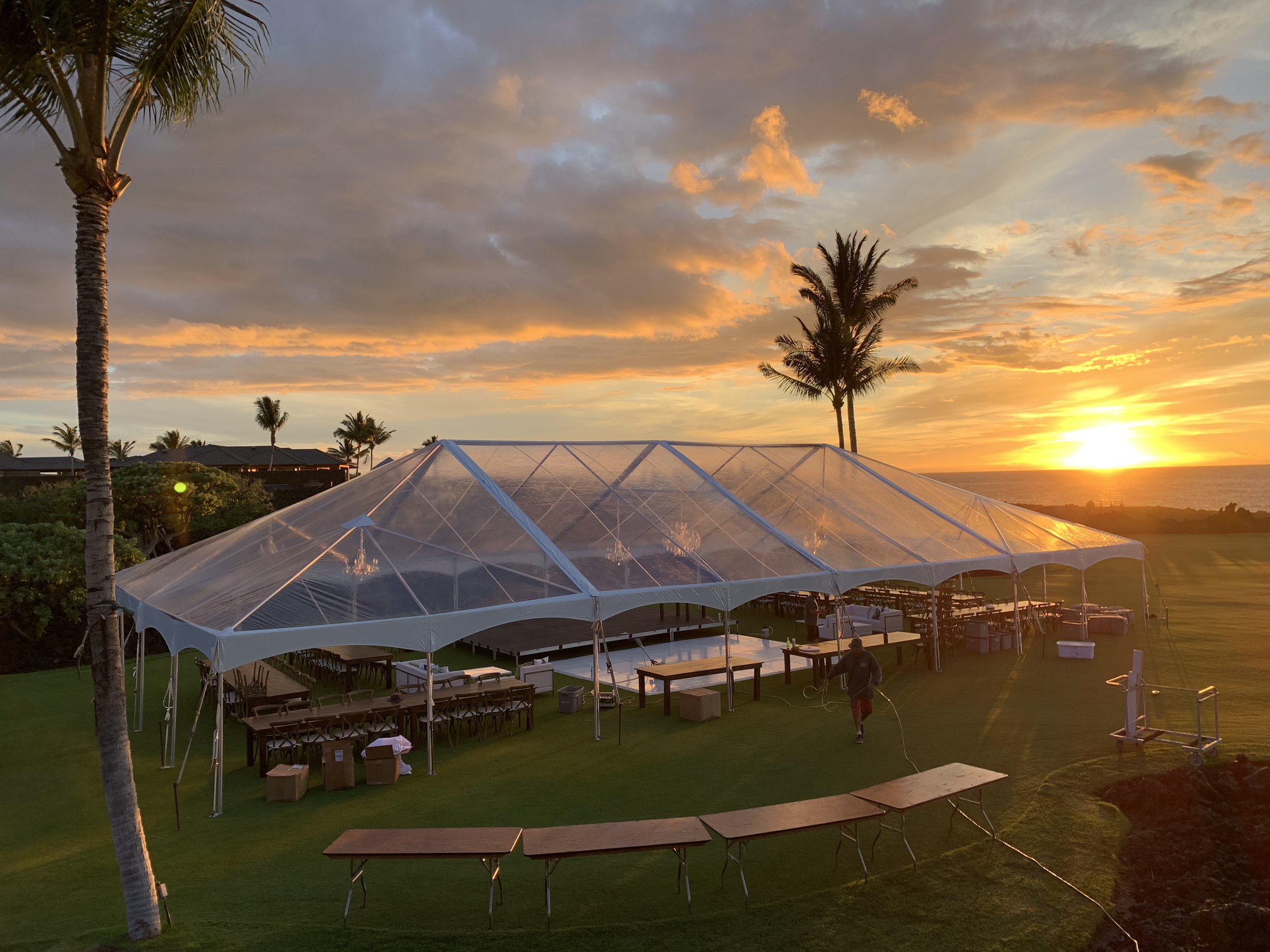 40x80 clear tent with white trim for wedding at Kukio North development. Featured in People Magazine. With Dark wood farm tables, crossback vineyard chairs, high gloss white dance floor, staging, chandeliers, and white leather lounge furniture.