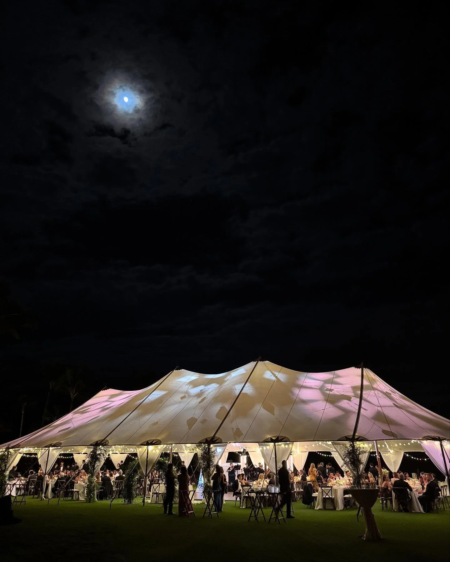 Beautiful wedding at Kaupulehu beach club featuring our 40x100 sail cloth tent, twinkle lighting with bamboo lanterns, white leg drapes, cross bank chairs, umbrellas, tall dance floor, white bars, staging, gold flatware, glass chargers, and tons of o