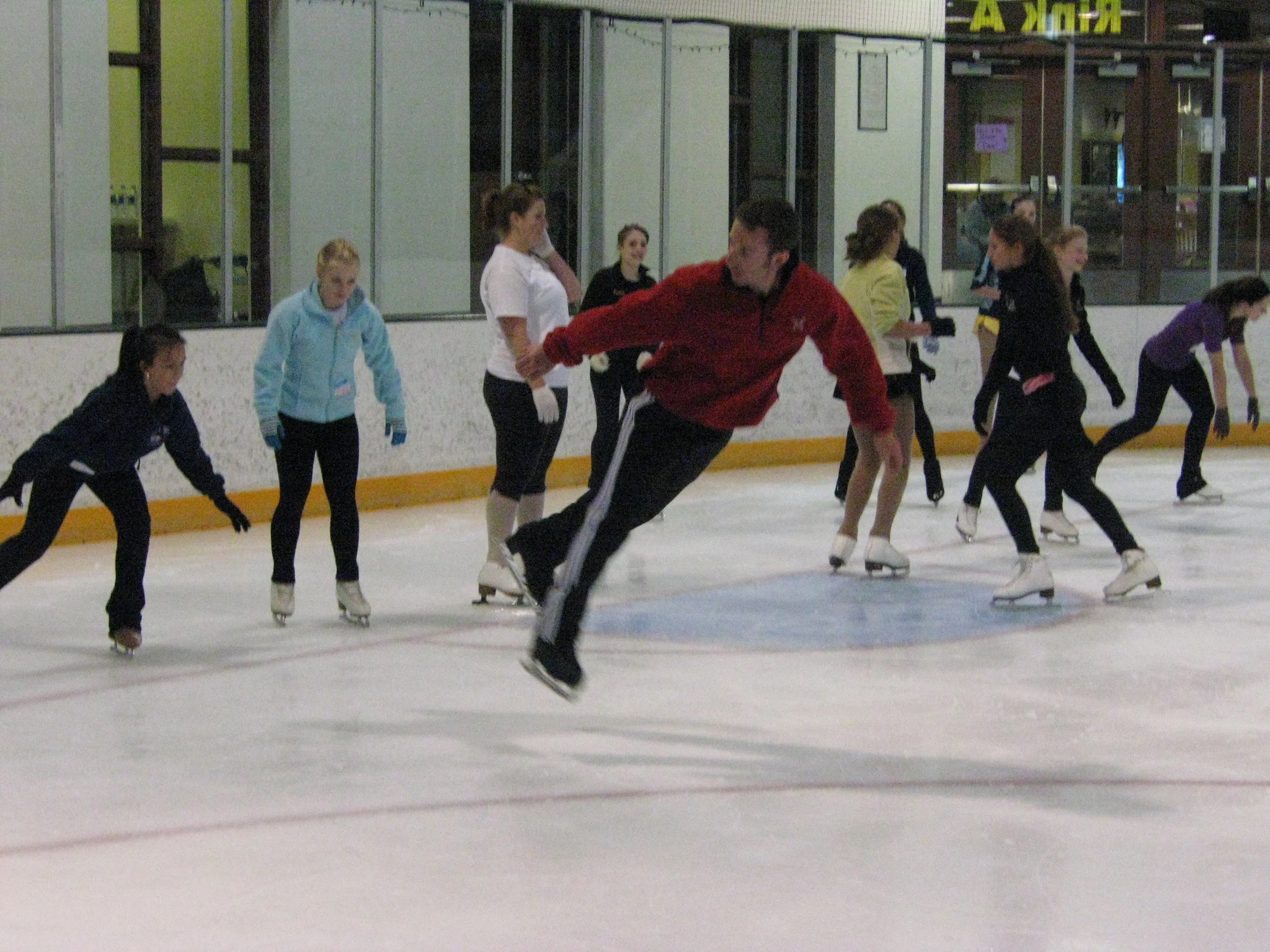 Group of people ice skating at an indoor ice rink, some skating and some observing.