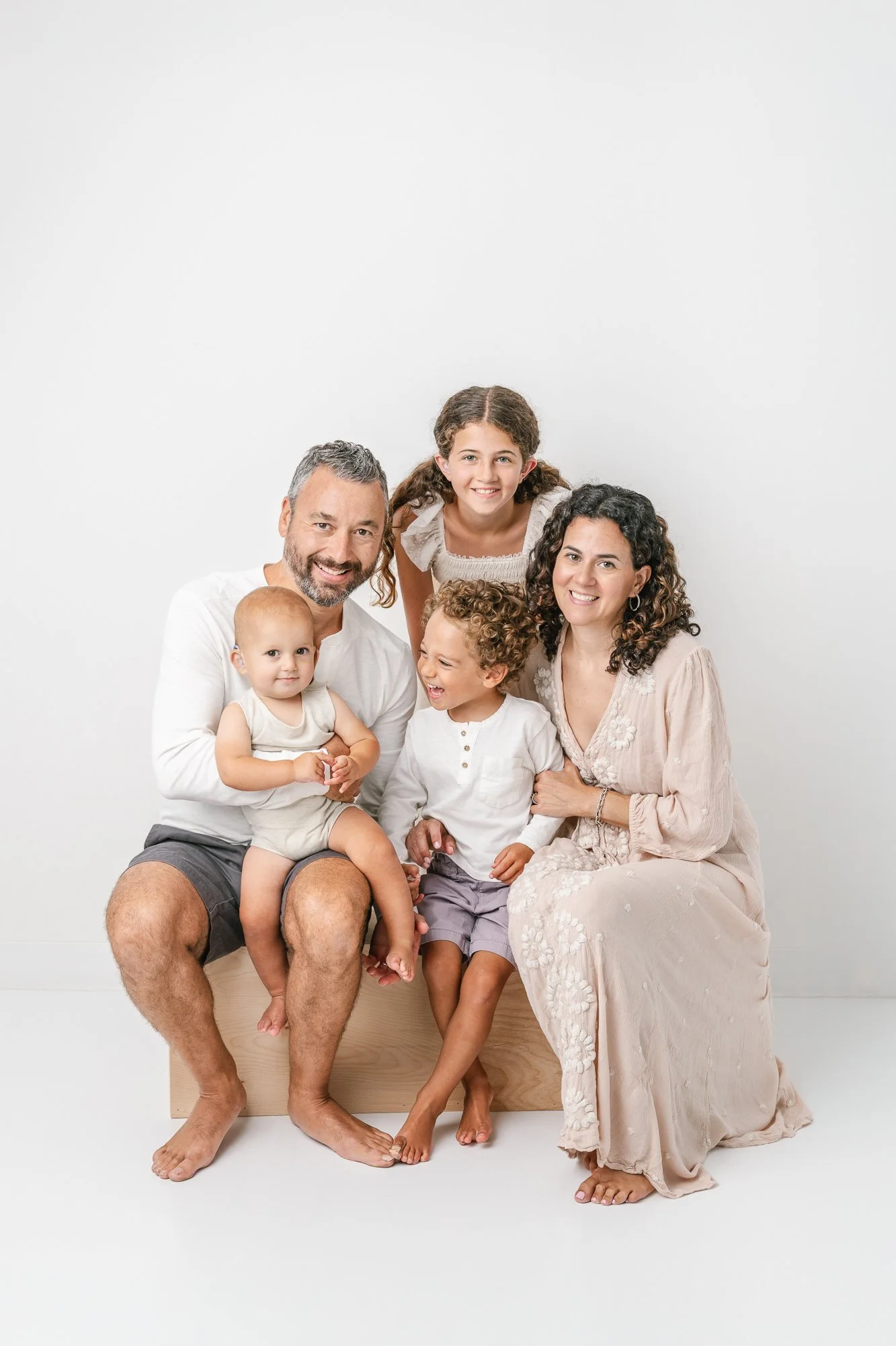 Smiling and casually dressed studio portrait of family of five, including a teenager, toddler, and one year old baby, sitting together against a white wall.