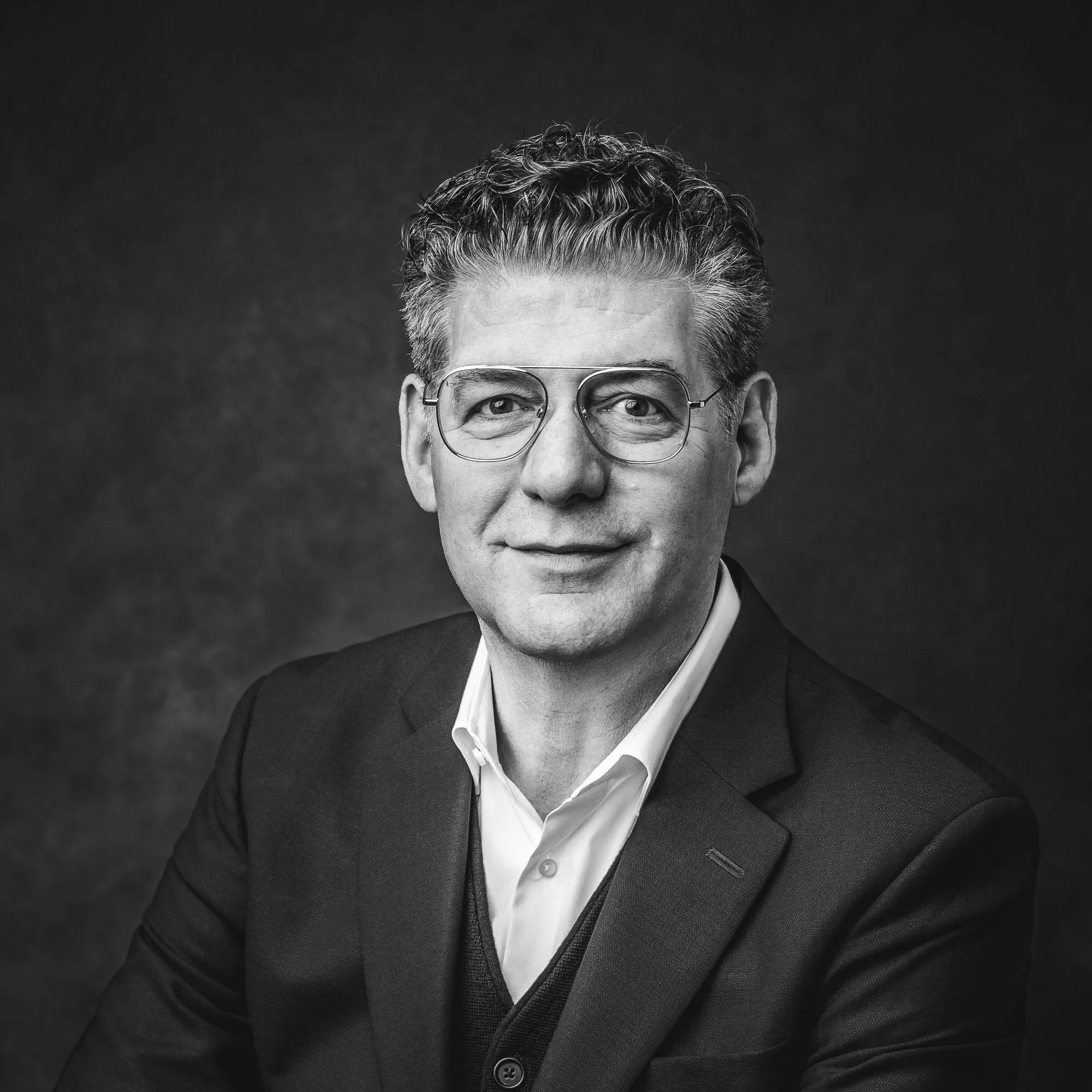 Black and white portrait of a professional man wearing glasses, a blazer, and a white shirt, with a serious but warm expression, against a dark background.