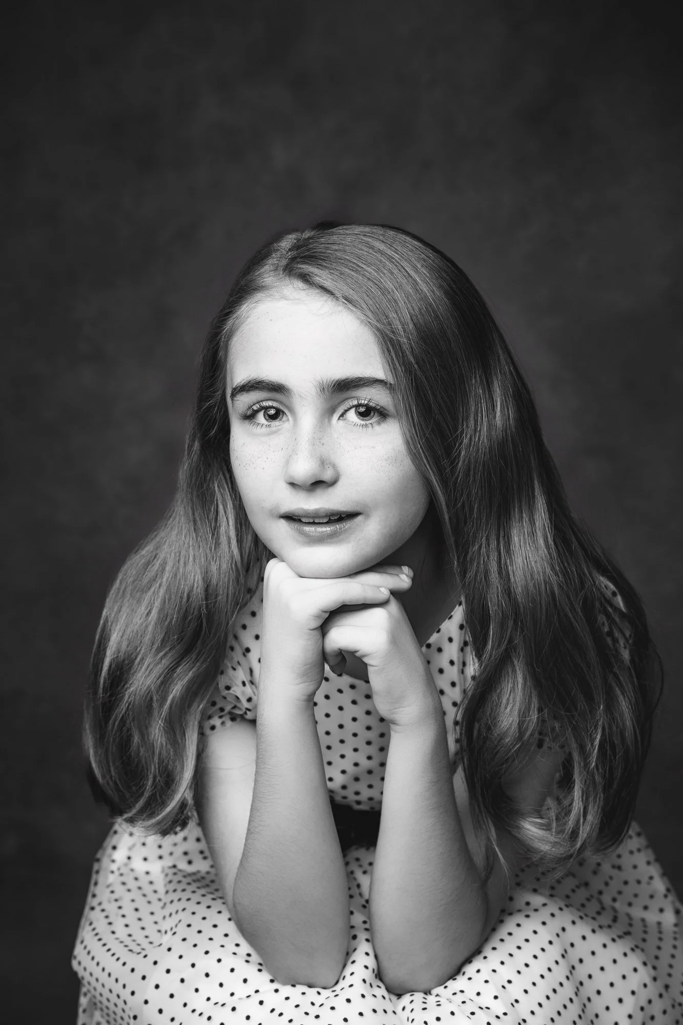 Black and white studio child portrait of a young girl with long hair, looking at the camera with a soulful expression and resting her chin on her hands.