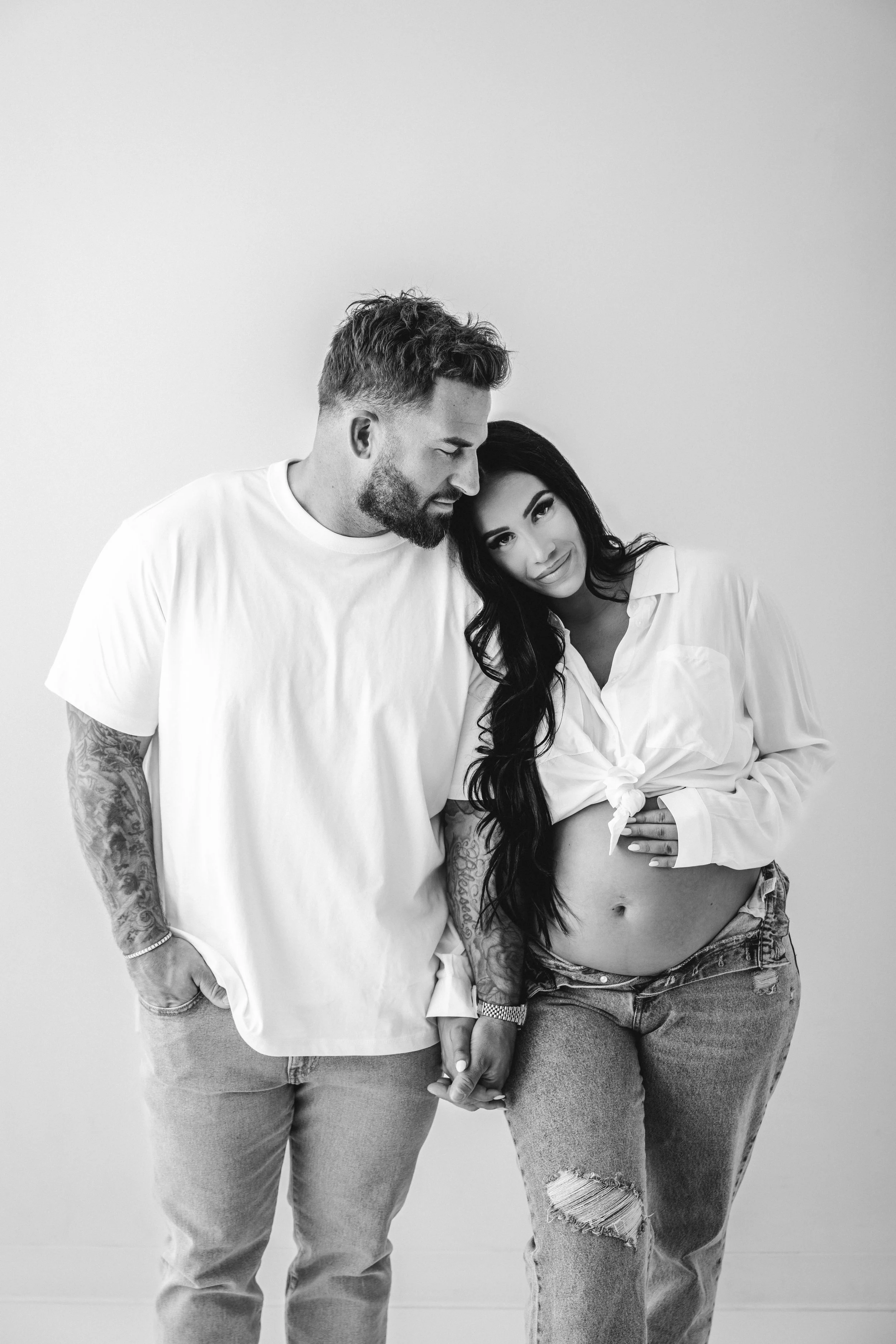 A modern black-and-white studio maternity portrait of a couple holding hands, with the woman gently touching her belly and husband kissing her head. Editorial maternity portrait vibe.