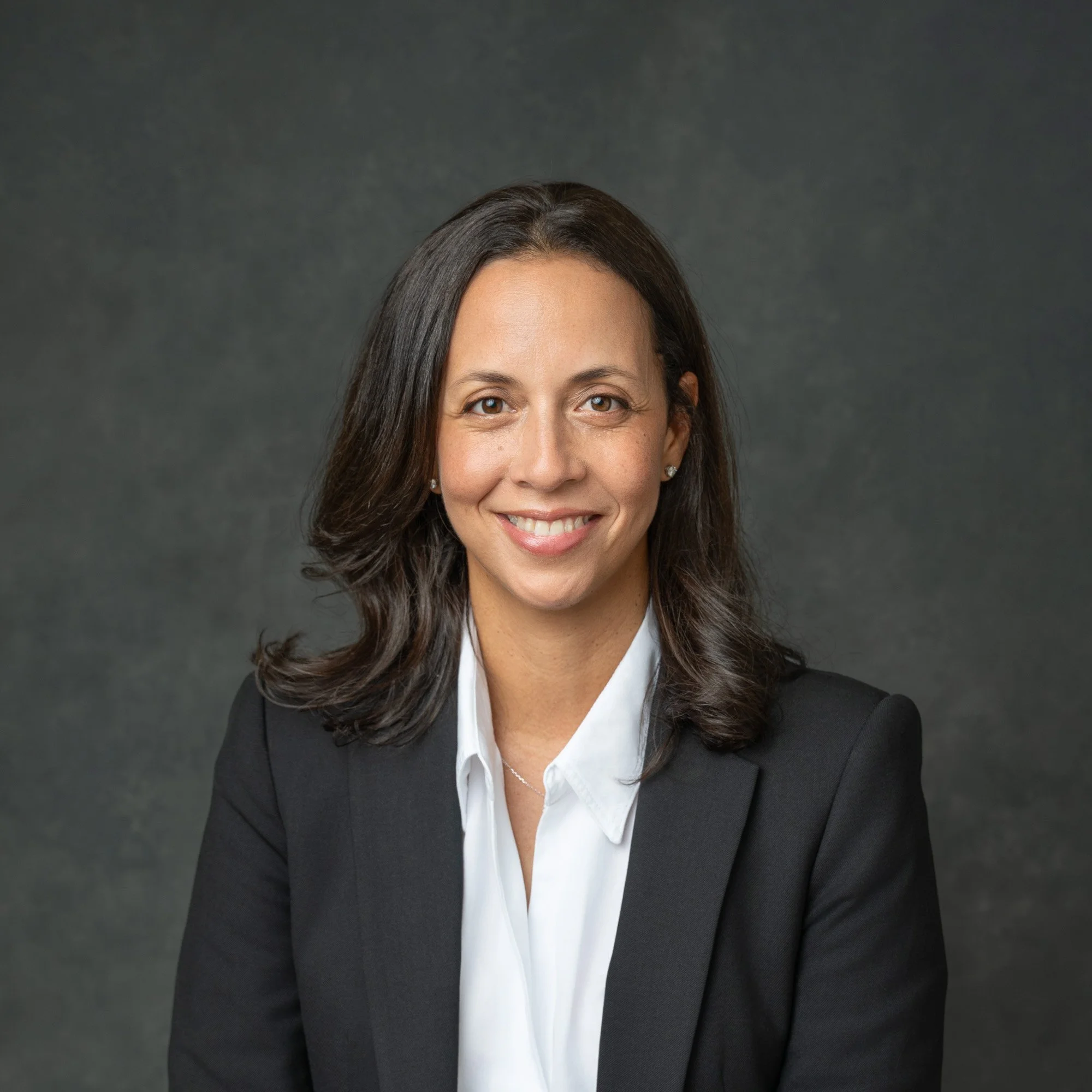 Professional portrait of a woman with brown hair wearing a black blazer and white shirt against a dark gray background.