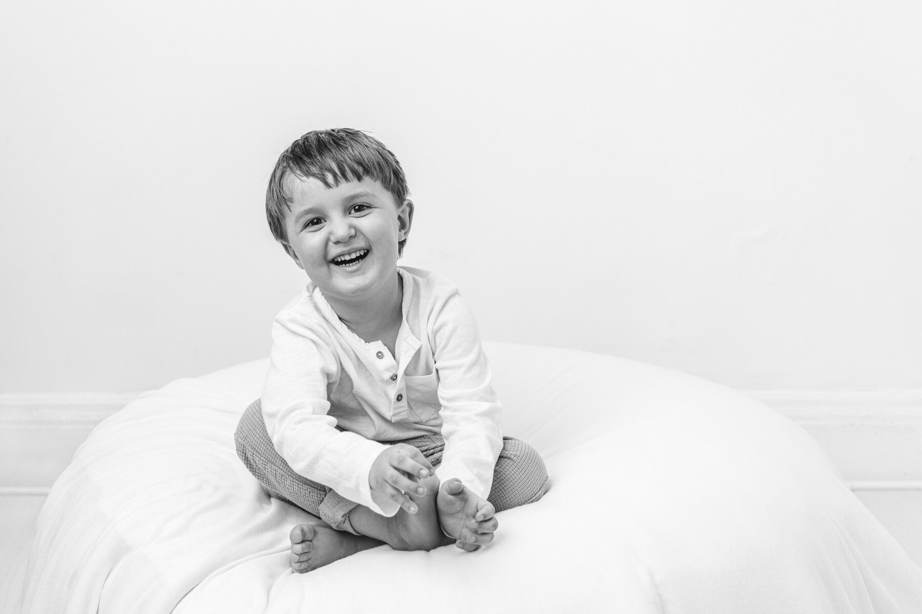 Happy_toddler_big_brother_sitting_on_newborn_beanbag_in_photography_studio_black_and_white