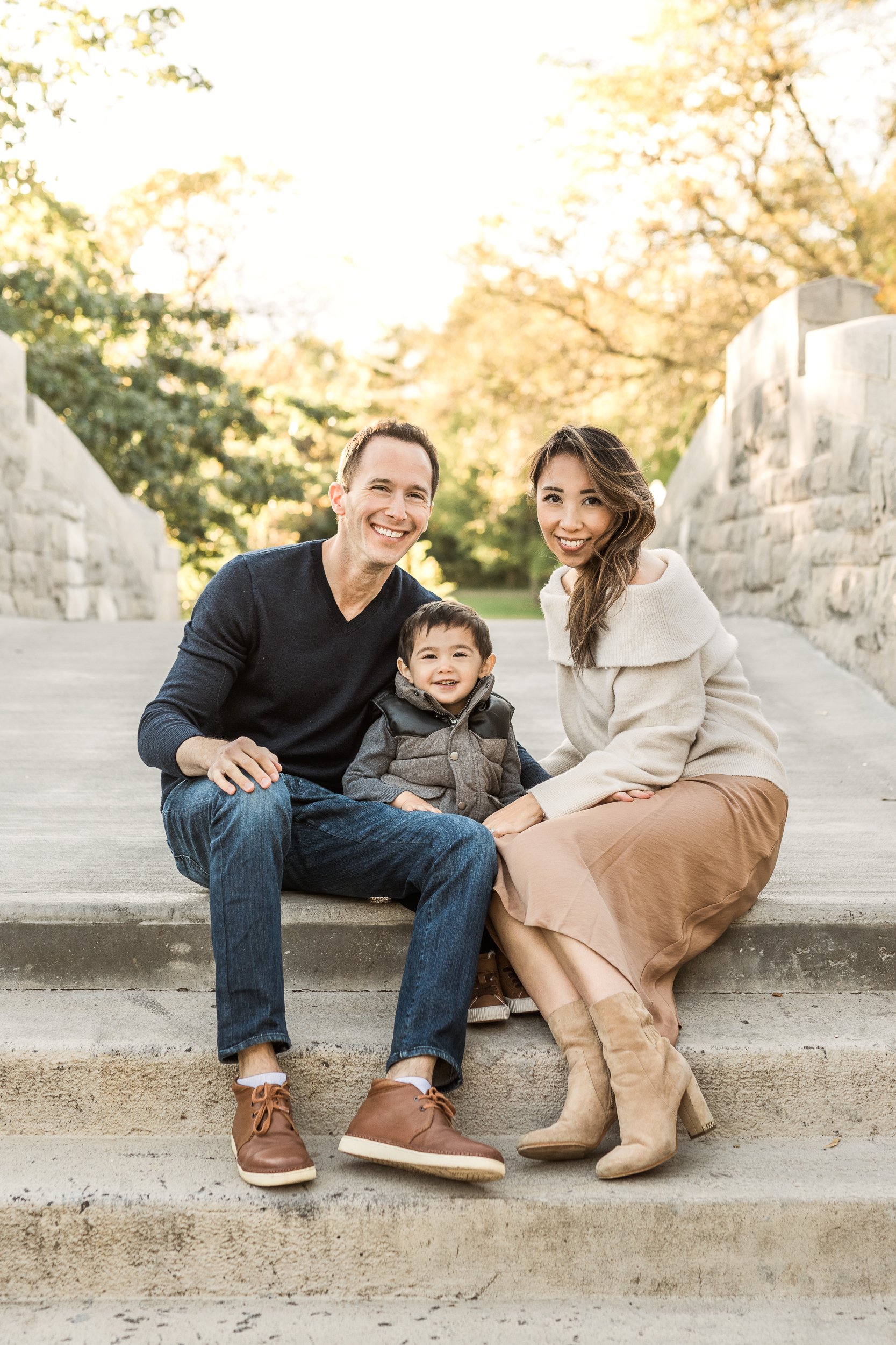  A young family wearing fall neutrals sit on cement steps and smile for a family portrait during a New Jersey Family session with Nicole Hawkins Photography. family portraits fall family attire new Jersey family photos #nicolehawkinsphotography #nico