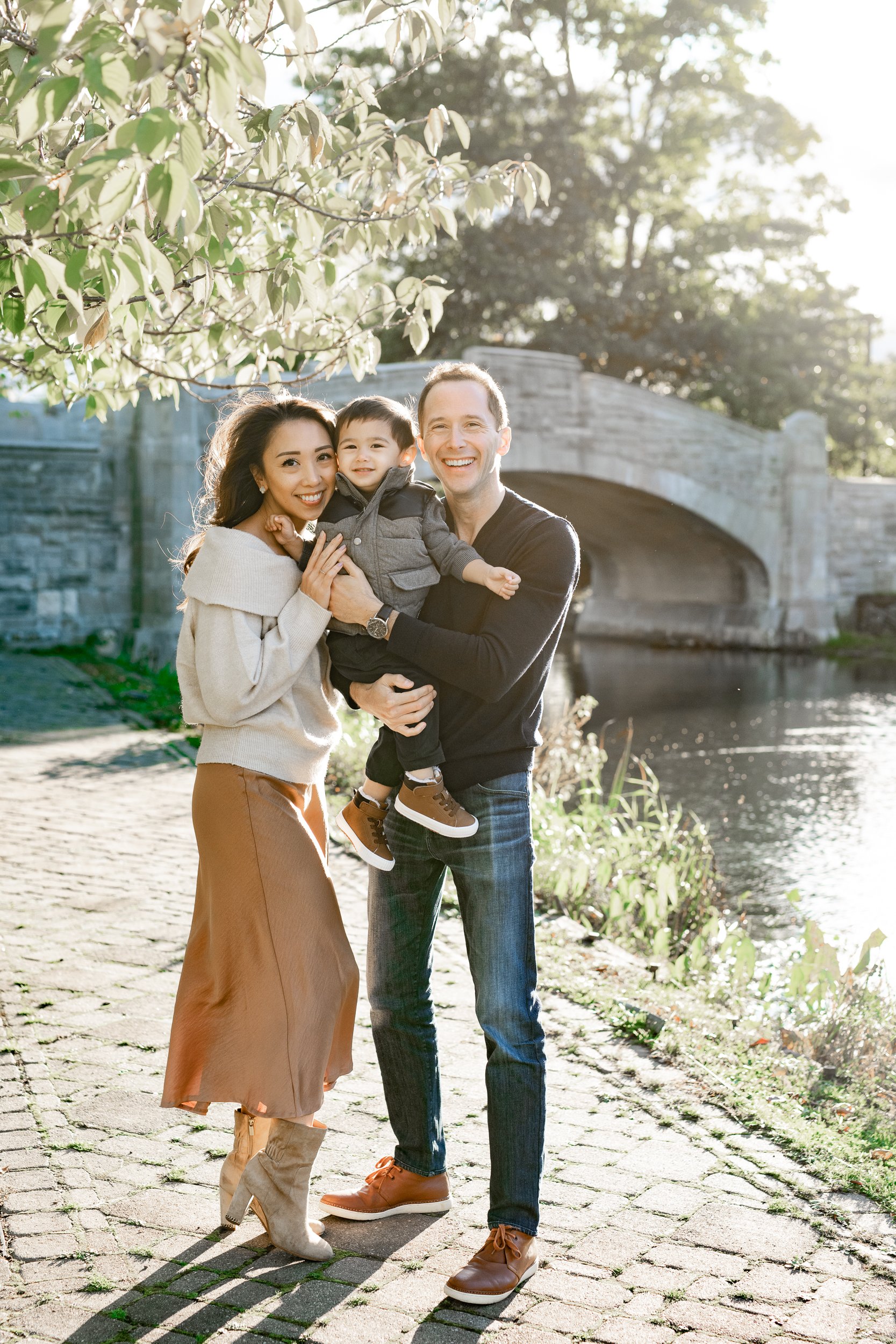  In Verona Park, Nicole Hawkins Photography captures a portrait of a darling family next to a river with golden sunlight highlights. cobblestone path pictures by the river stone bridge young family #nicolehawkinsphotography #nicolehawkinsfamilies #ne