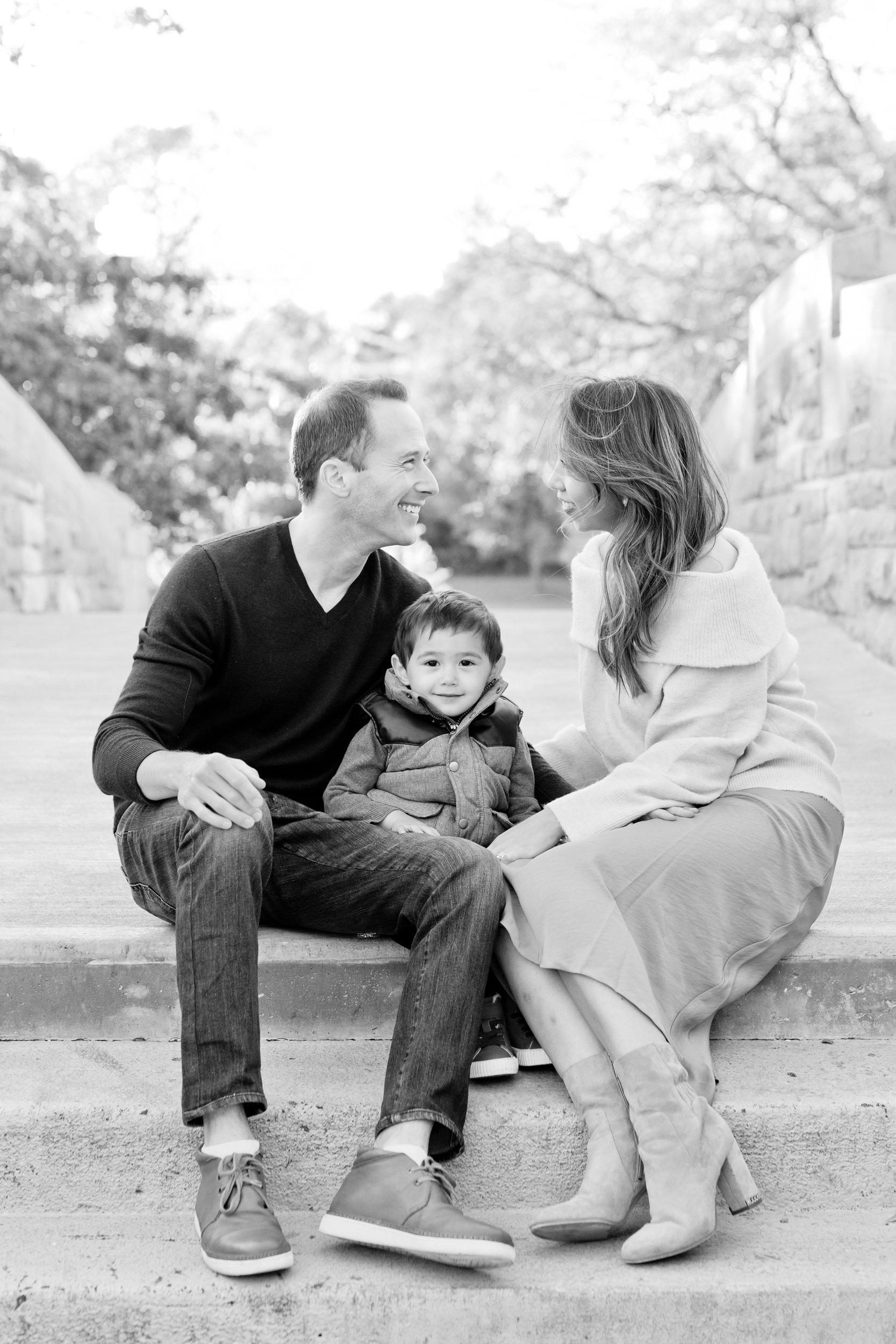  Black and white photo of a mother and father smiling at one another as their little boy looks forward by Nicole Hawkins Photography a New Jersey Family Photographer. one little boy black and white family pic mom in a skirt #nicolehawkinsphotography 