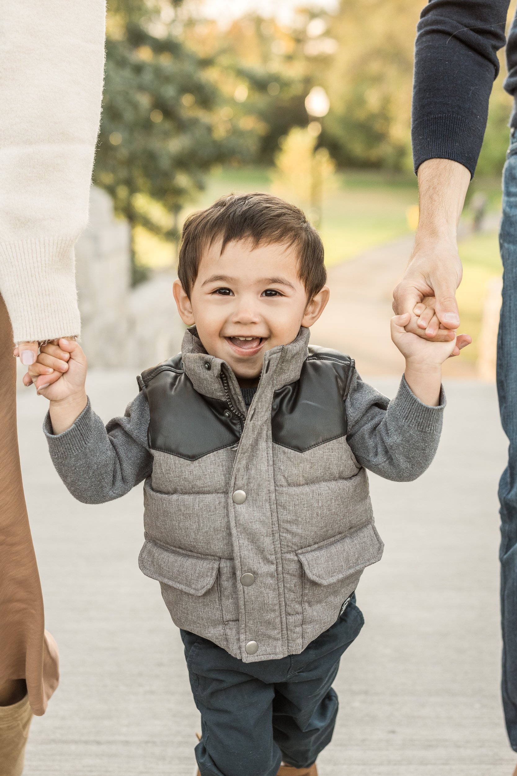  A smiling young boy with a gray sweater and long sleeve shirt holds on to his parent's hands to walk by Nicole Hawkins Photography of New Jersey. young family photographer new jersey family portraits #nicolehawkinsphotography #nicolehawkinsfamilies 