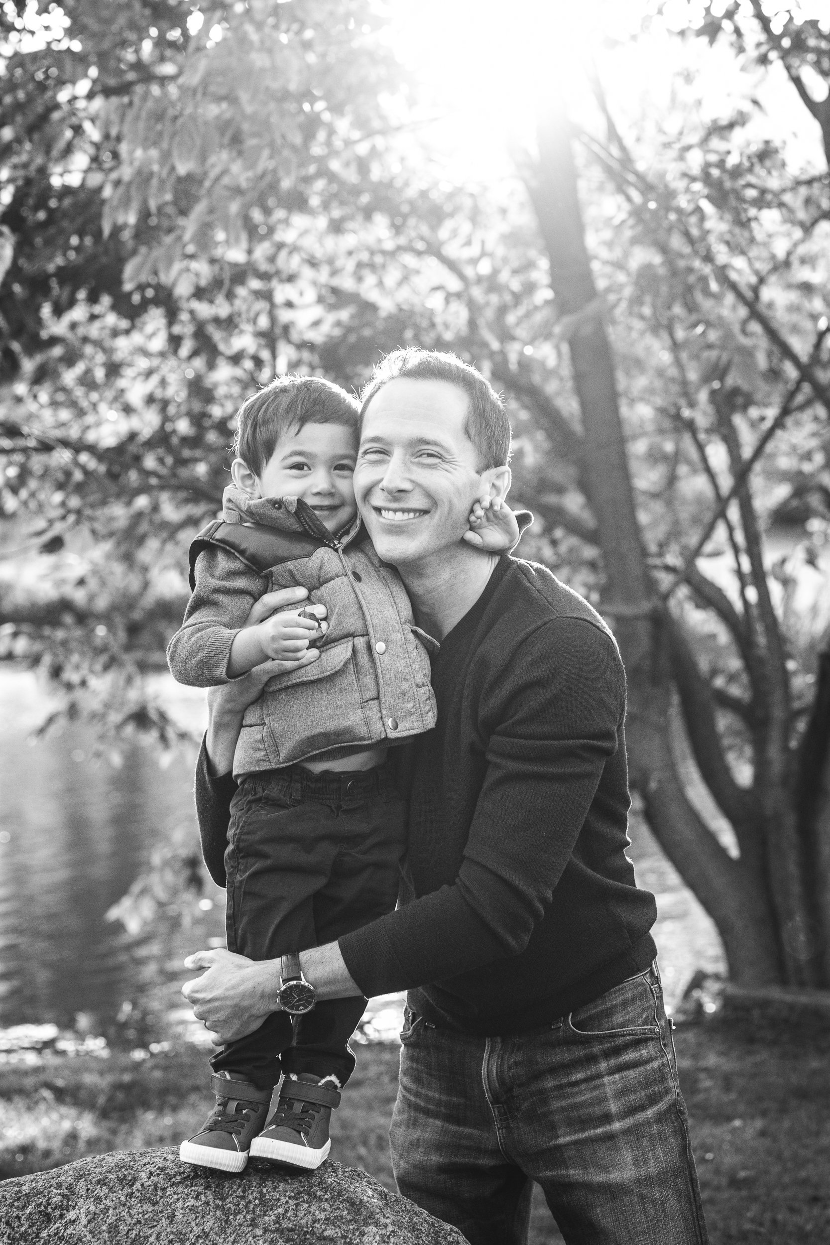  A toddler stands on a rock as his father hugs him tightly, both with big smiles on their face at Verona Park in New Jersey by Nicole Hawkins Photography. father and son pic dad and lad portraits big smiles with daddy #nicolehawkinsphotography #nicol