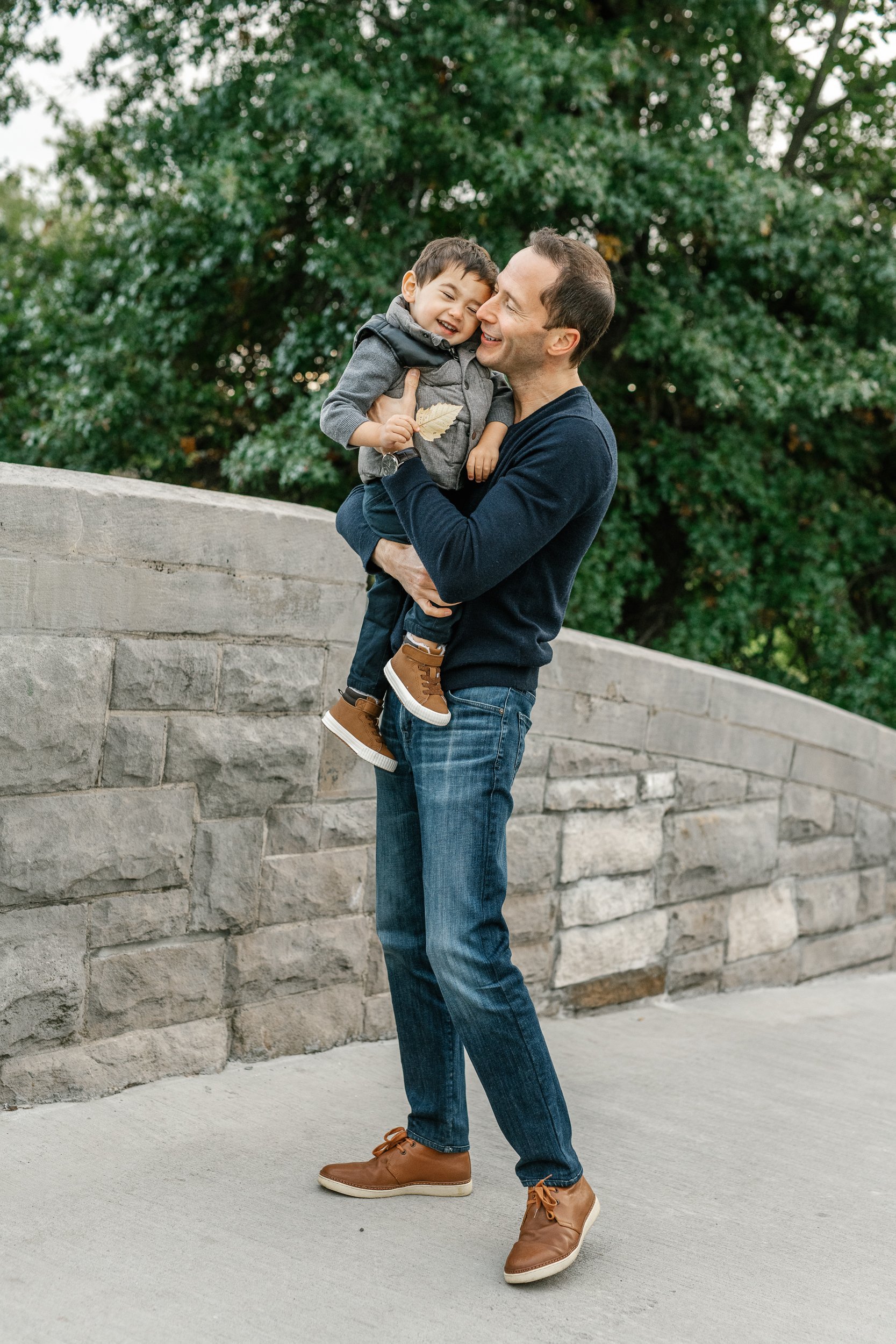 A father holds his little boy in his arms and tickles him in Verona Park on a stone bridge captured by Nicole Hawkins a family photographer in New Jersey. brown dress shoes for men brown boots boys father and son portrait #nicolehawkinsphotography #