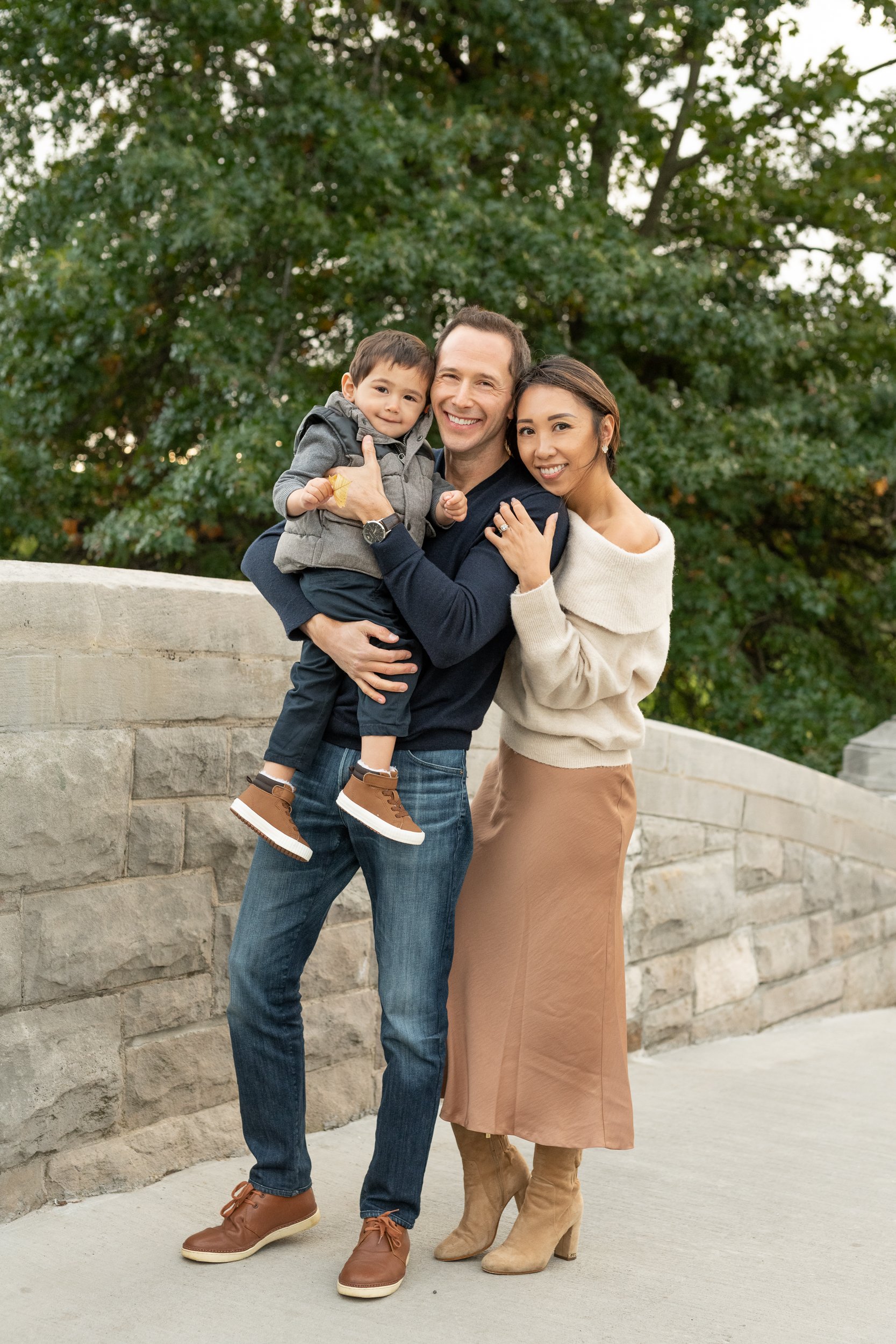  A young family squishes together in a New Jersey Park in the fall wearing neutral colors captured by Professional photographer Nicole Hawkins Photography. family fall outfit inspiration women's fall sweater sweater with skirt #nicolehawkinsphotograp