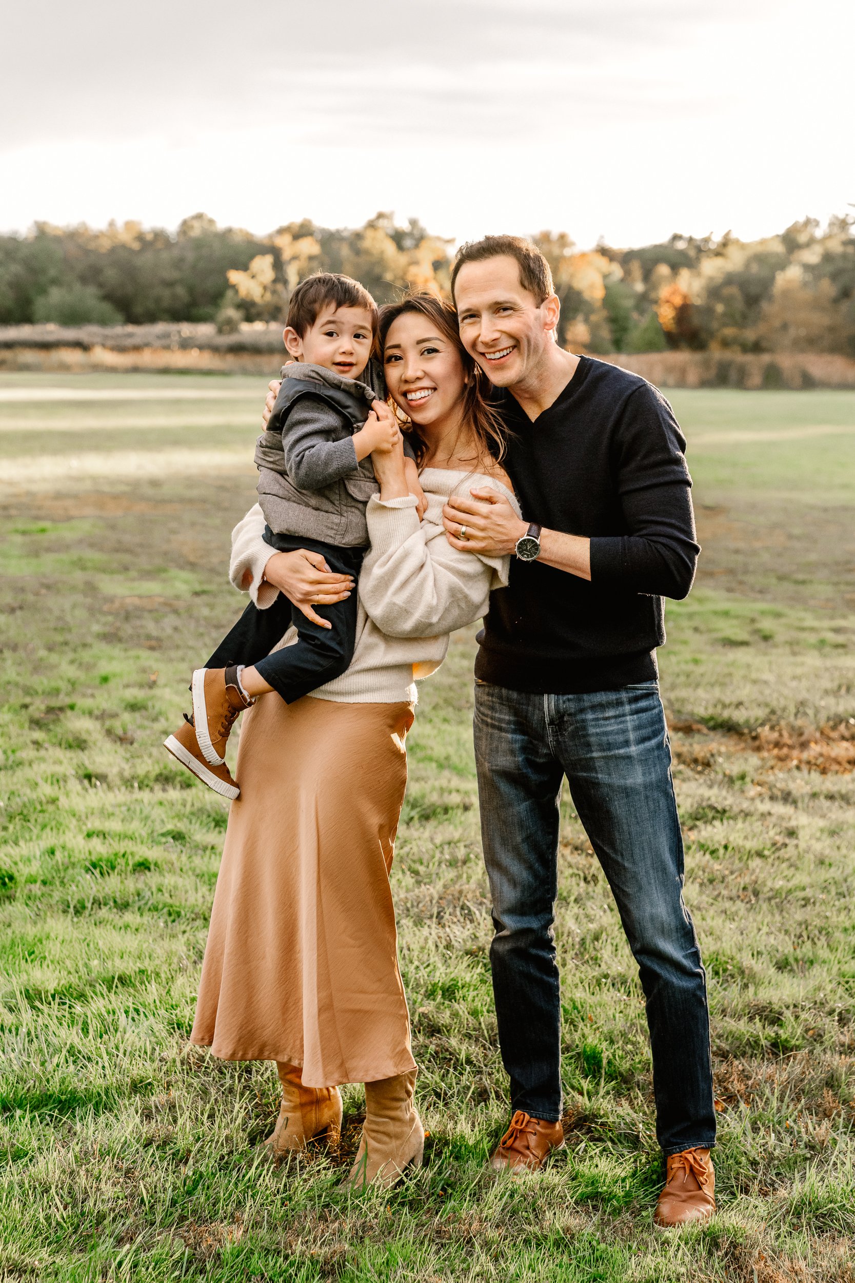  In a large green grass field with fall leaves in the background a family of three smiles while the sunsets in Verona Park by Nicole Hawkins Photography. sunset family photos NJ sunset family photographers Verona park family photos #nicolehawkinsphot