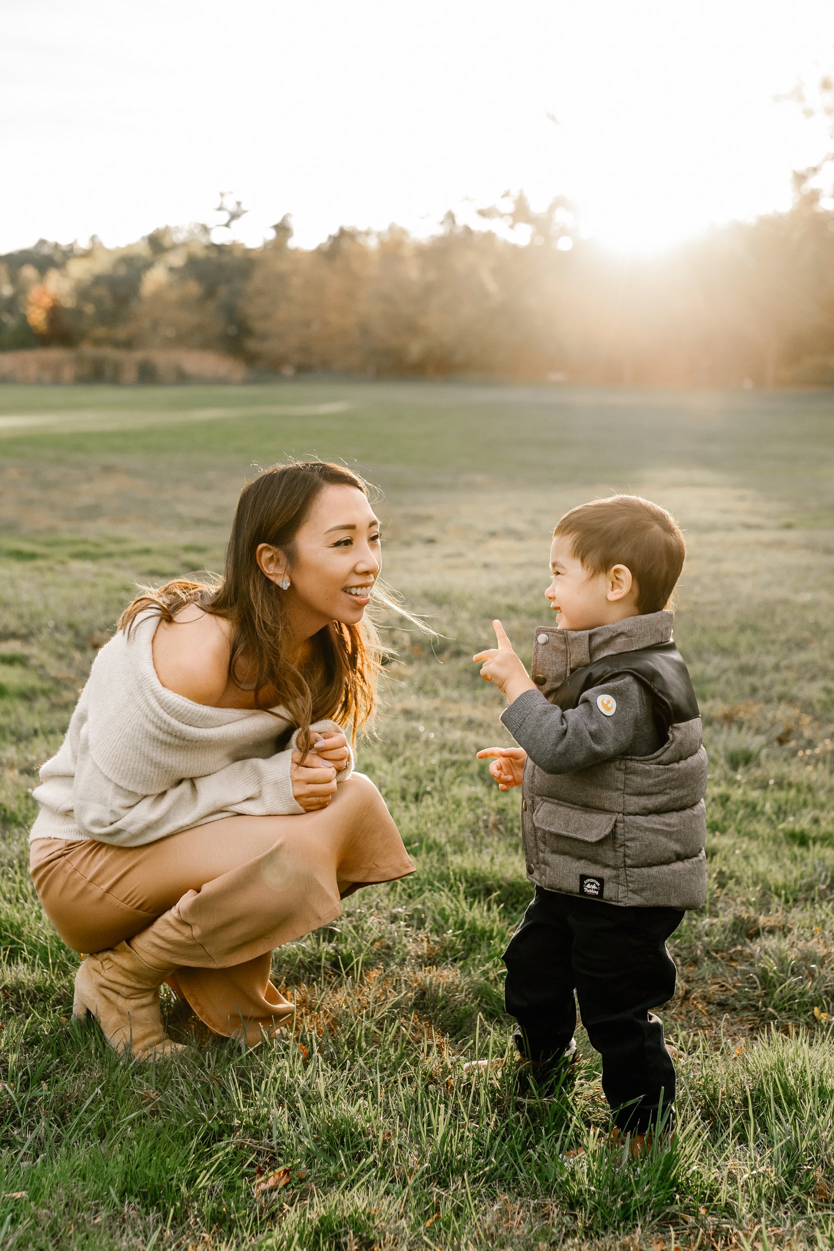  A mother in a cream sweater and tan skirt bends down and plays with her toddler little boy during a family session with Nicole Hawkins Photography in NJ. tan skirt with sweater gray toddler shirt playing pictures family goals #nicolehawkinsphotograp