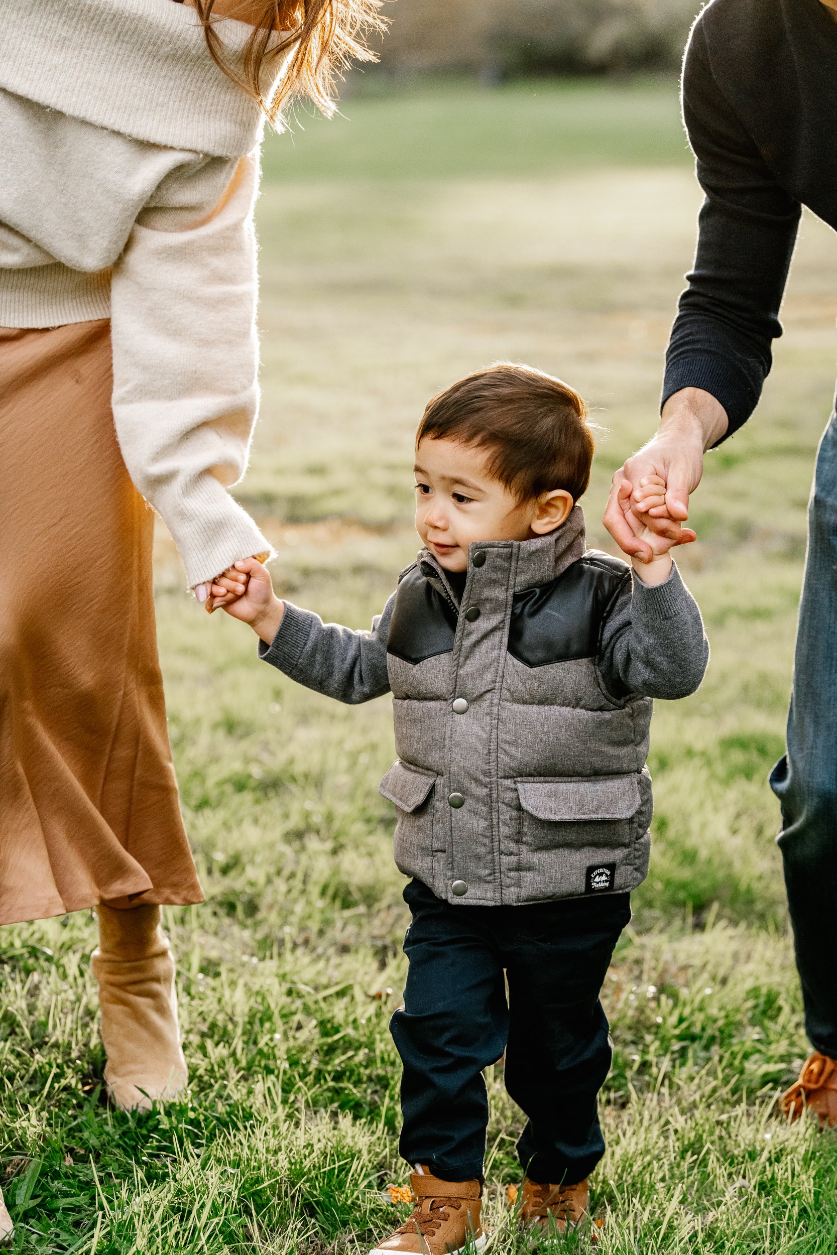 During fall time in New Jersey, a young boy wearing black and gray holds his parent's hands in a green grass field by Nicole Hawkins Photography. boy in black and gray green grass field outdoor New Jersey family pictures #nicolehawkinsphotography #n