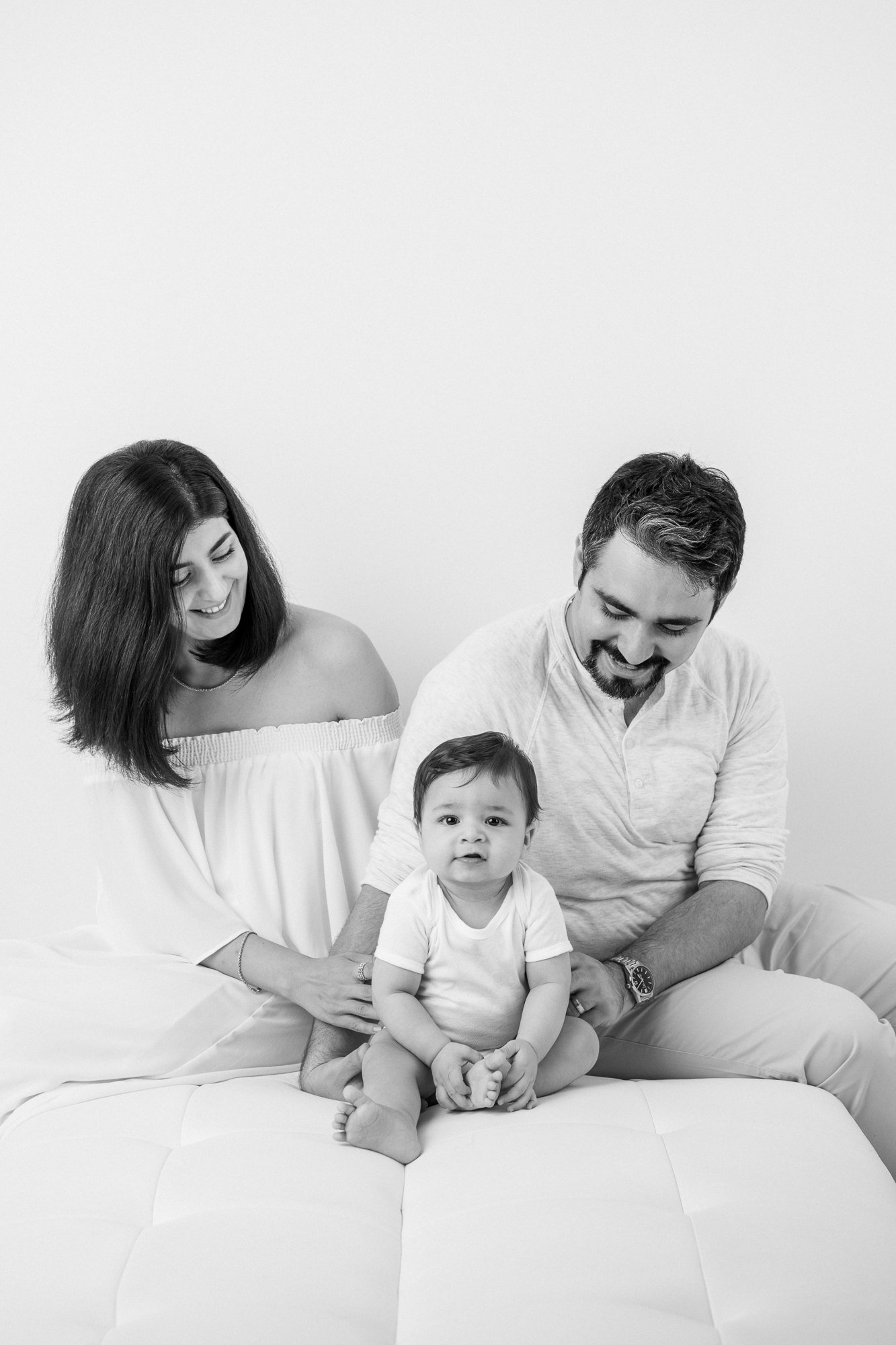  Mother and father sit behind their baby boy and smile sweetly down at him in a bright New Jersey studio by Nicole Hawkins Photography. family of three first born baby baby boy with parents in a studio #nicolehawkinsphotography #nicolehawkinsbabies #