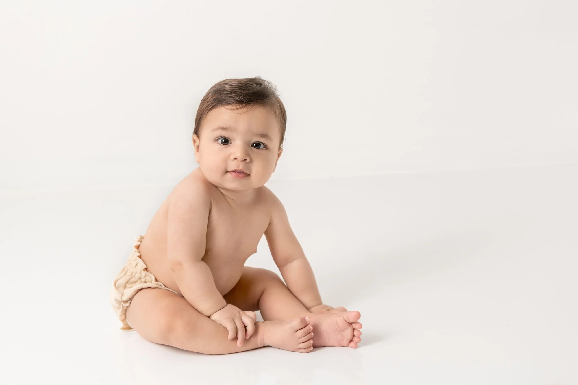  With a tan diaper on a little boy sits up with his hand on his chunky legs and looks up with a white studio background by New Jersey baby photographer Nicole Hawkins Photography. New Jersey child photographer NJ milestone photographer #nicolehawkins