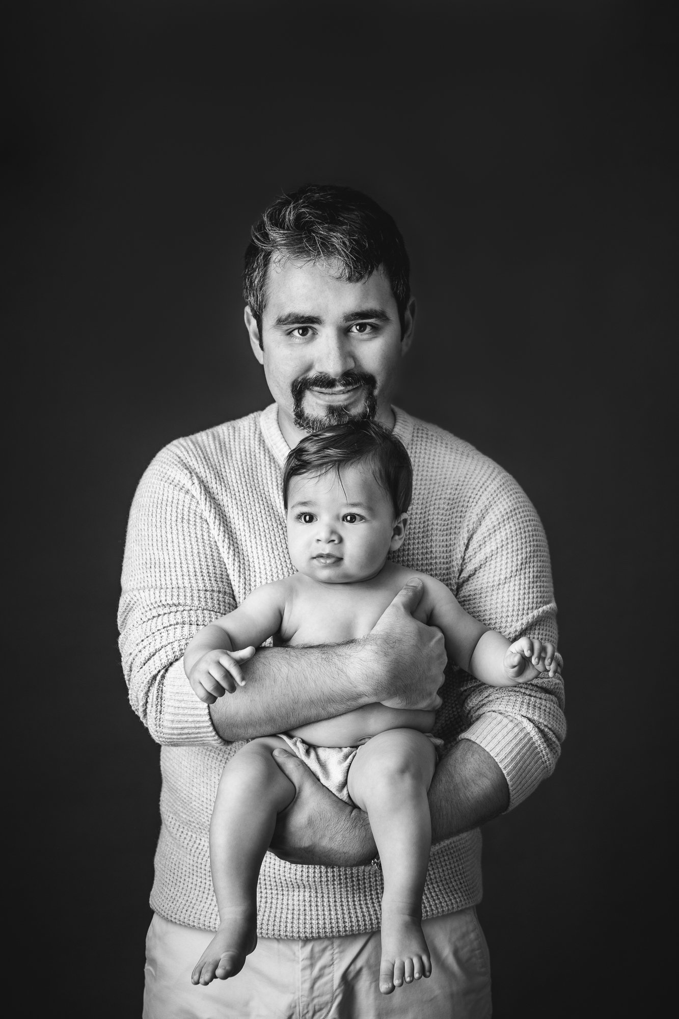  A New Jersey baby photographer captures a black and white portrait of a father holding a baby boy in his arms and looking forward by Nicole Hawkins Photography. stunning black and white father and baby portrait black background shot #nicolehawkinsph