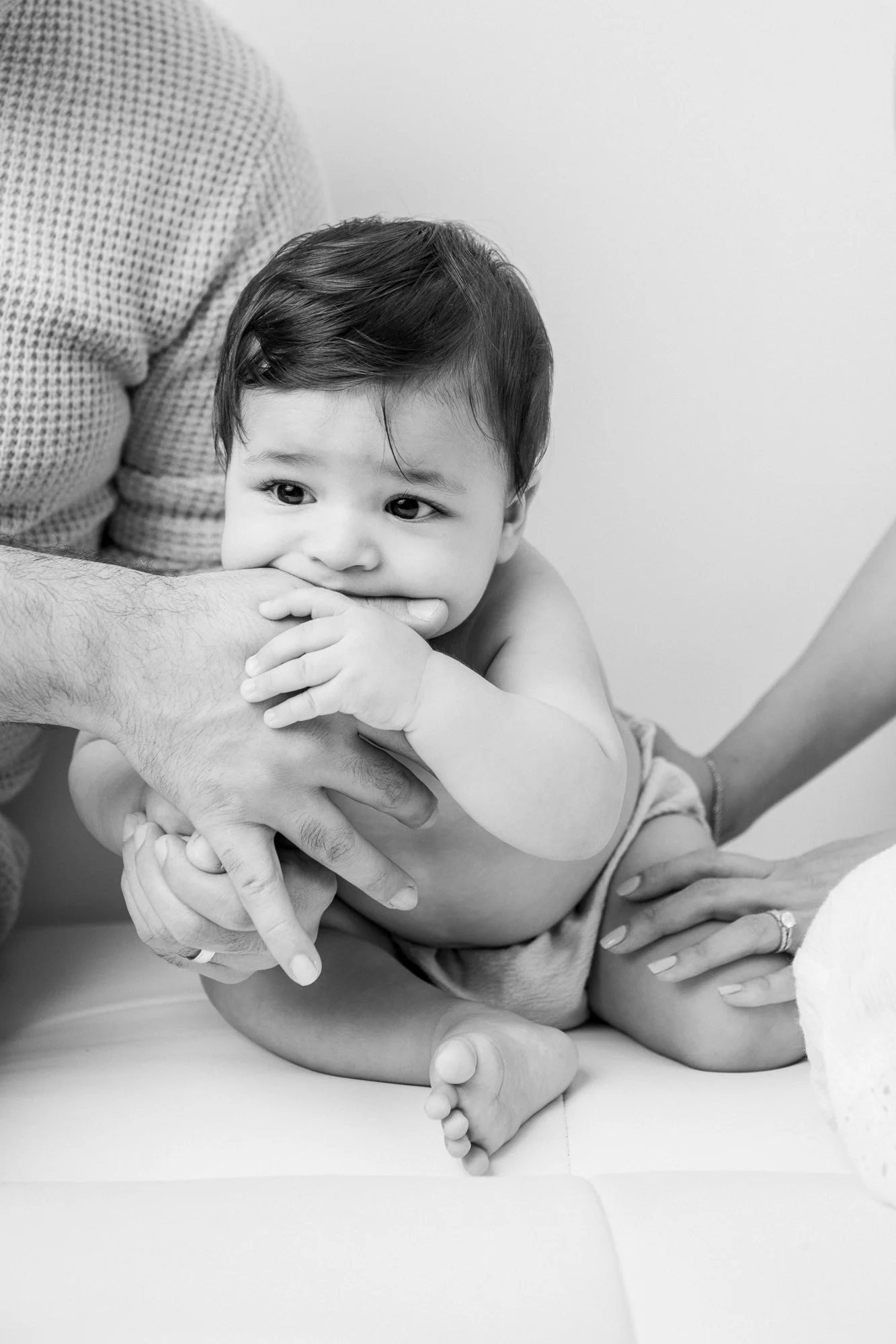  A little boy sucks his dad's hand while his mother holds him up during a New Jersey Studio Portrait session with Nicole Hawkins Photography. baby sucking on dad's hand NJ studio family portraits baby boy #nicolehawkinsphotography #nicolehawkinsbabie