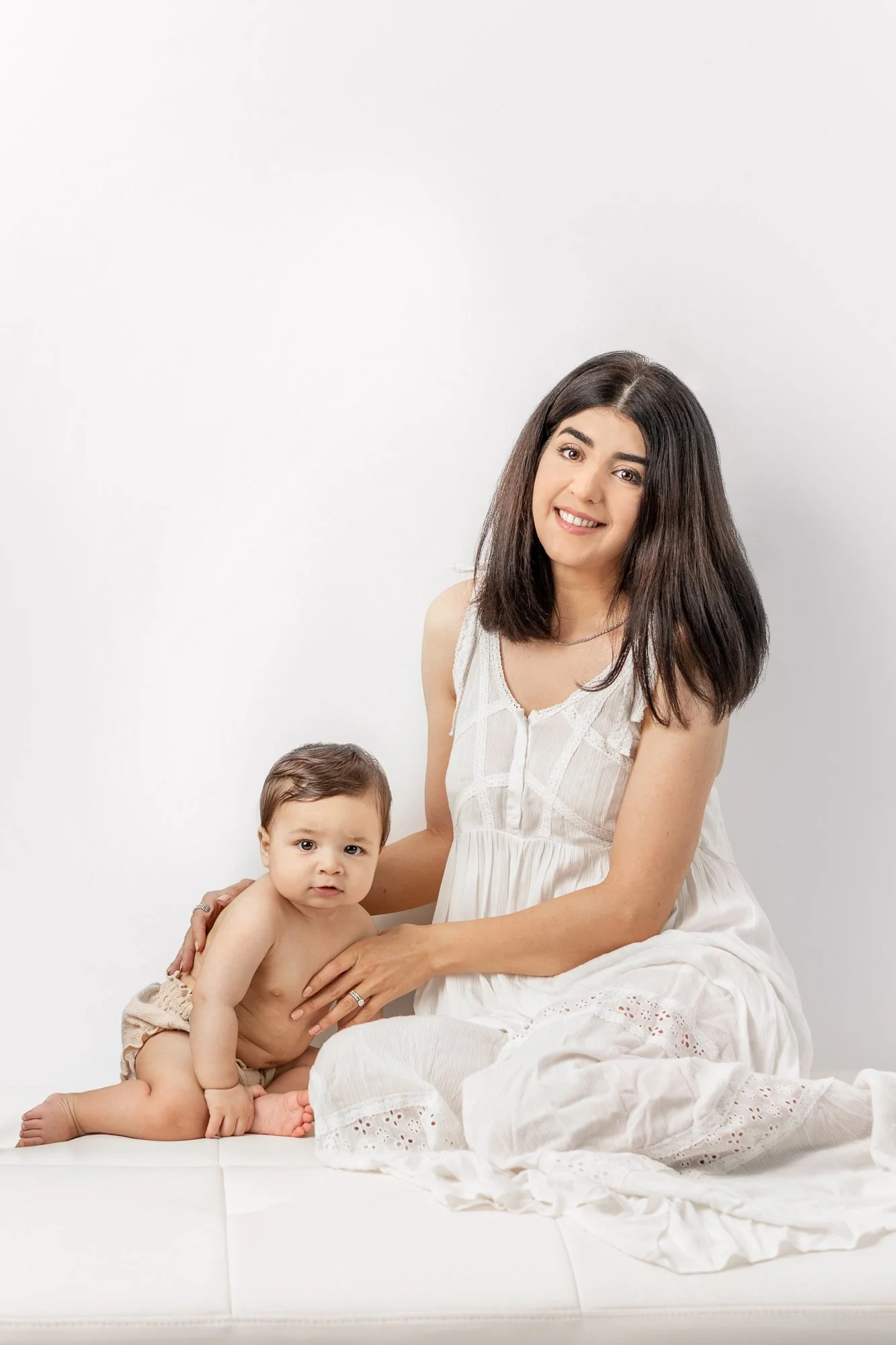  A mother wearing a white dress sits next to her baby boy and looks forward at a Studio in New Jersey by Nicole Hawkins Photography. mother and baby mother in white all white in a studio simple studio attire #nicolehawkinsphotography #nicolehawkinsba