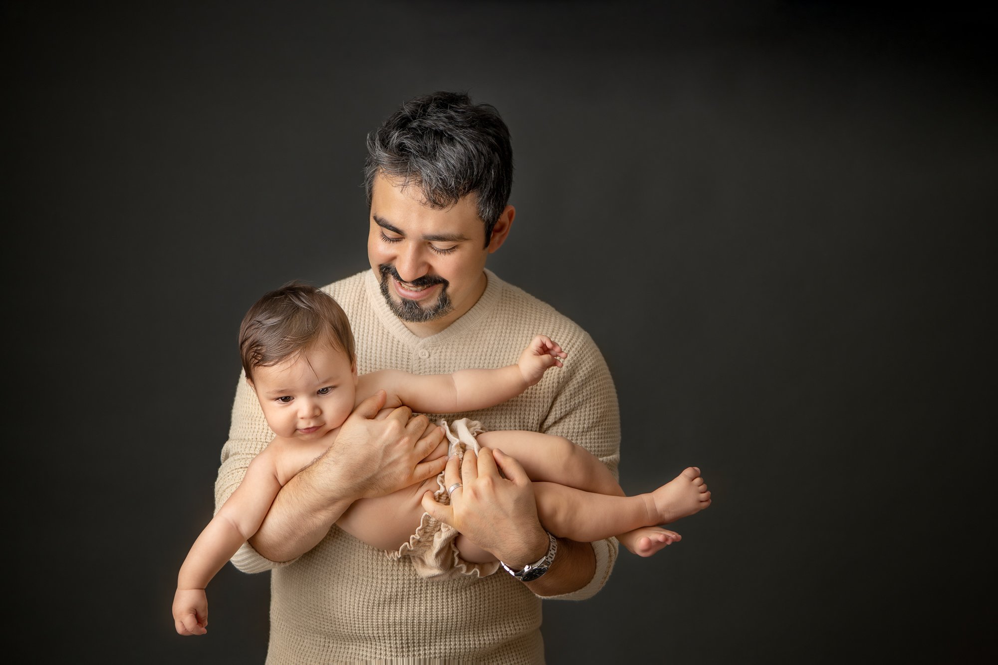  With a black background in a New Jersey studio, a father plays with his young son captured by Nicole Hawkins Photography. timeless family photos father and son portrait in studio father plays with baby boy #nicolehawkinsphotography #nicolehawkinsbab