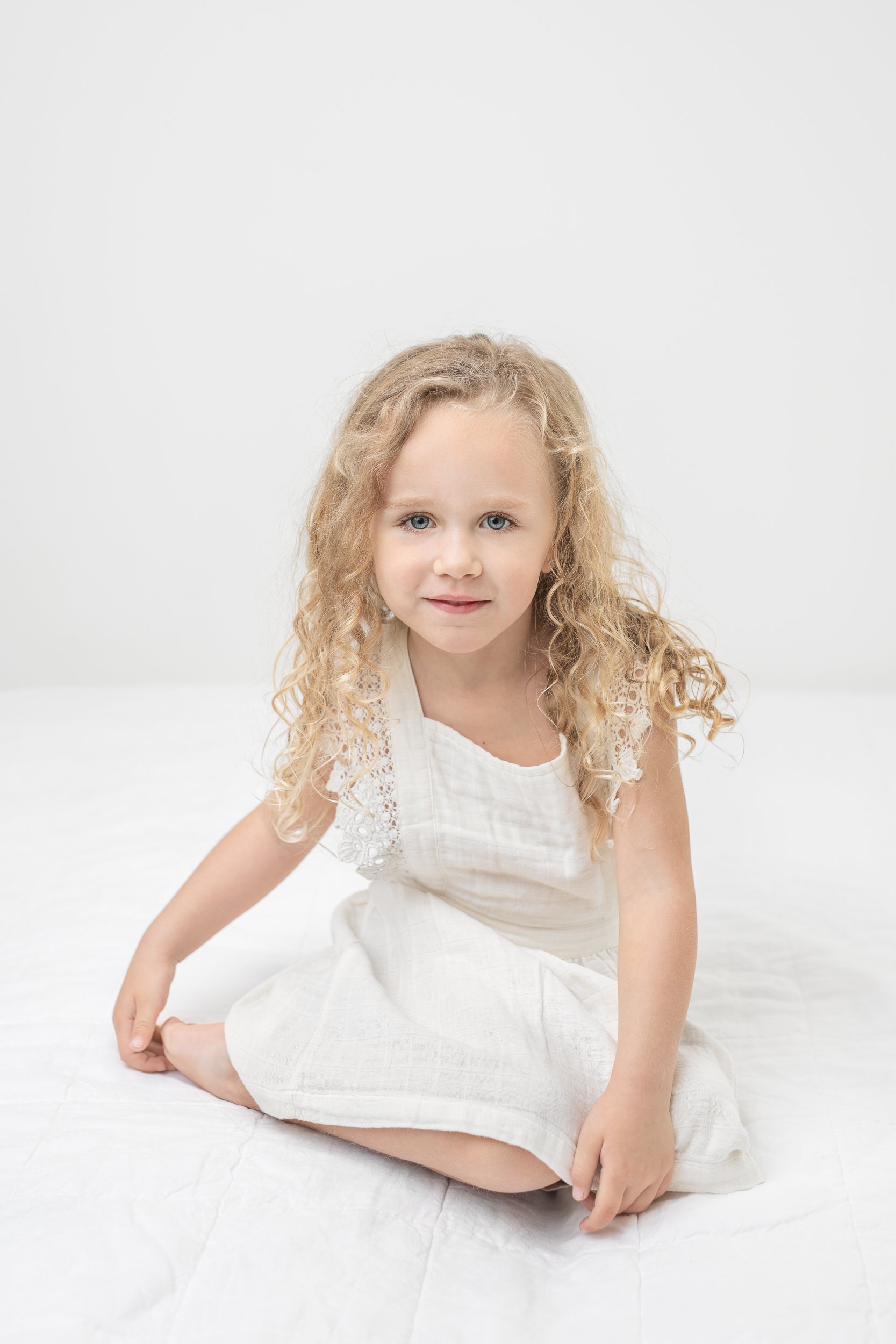  Blonde curly-haired little girl wearing a flutter sleeve white dress sitting on white bedding in a studio by Nicole Hawkins Photography a New Jersey photographer. little girl with blonde hair little girl studio session big sister girl outfit inspira