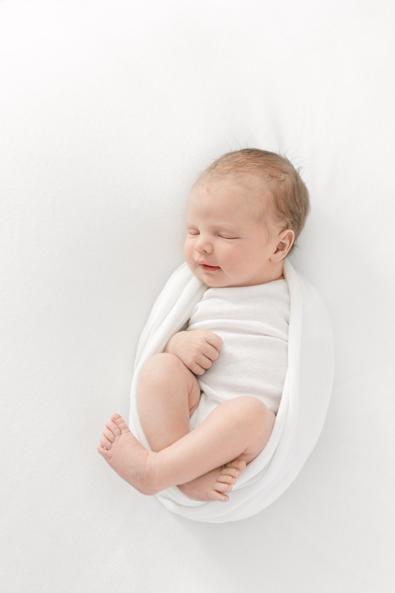  Tender newborn baby wrapped in a white swaddle with her legs sticking out and sleeping captured in a New Jersey studio by Nicole Hawkins Photography. baby wrapped in a swaddle all-white studio newborn session baby swaddle pose ideas baby feet baby l