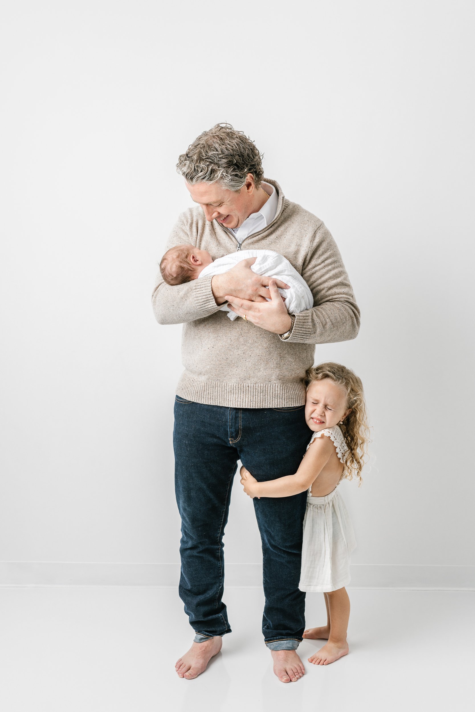  Father wearing a sweater holding his brand new baby girl with his older daughter holding onto his leg in New Jersey taken by Nicole Hawkins Photography. father and two daughters father and baby father and kids studio father outfit ideas #nicolehawki