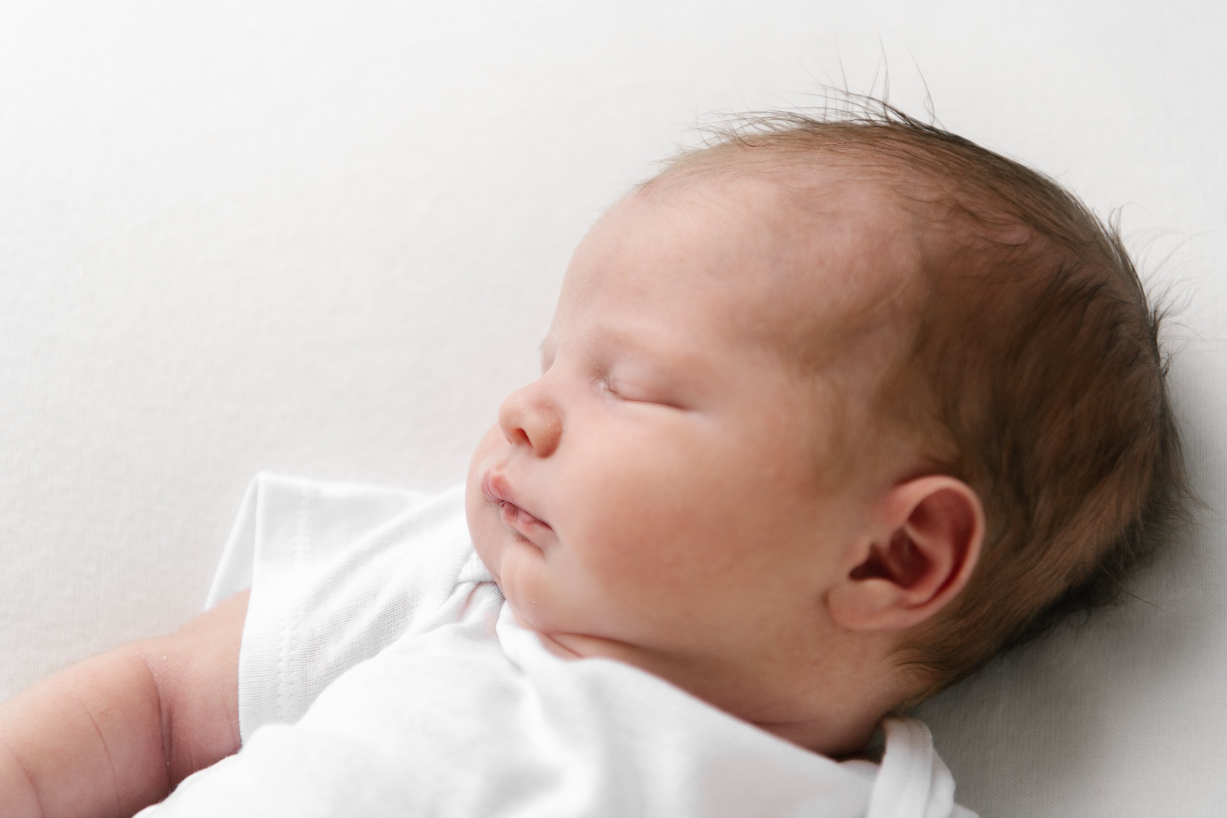  A baby with brown thin hair and a button nose sleeps peacefully during a studio newborn session in New Jersey with Nicole Hawkins Photography. baby with brown hair button nose baby white onesie on baby baby head close-up #nicolehawkinsphotography #n