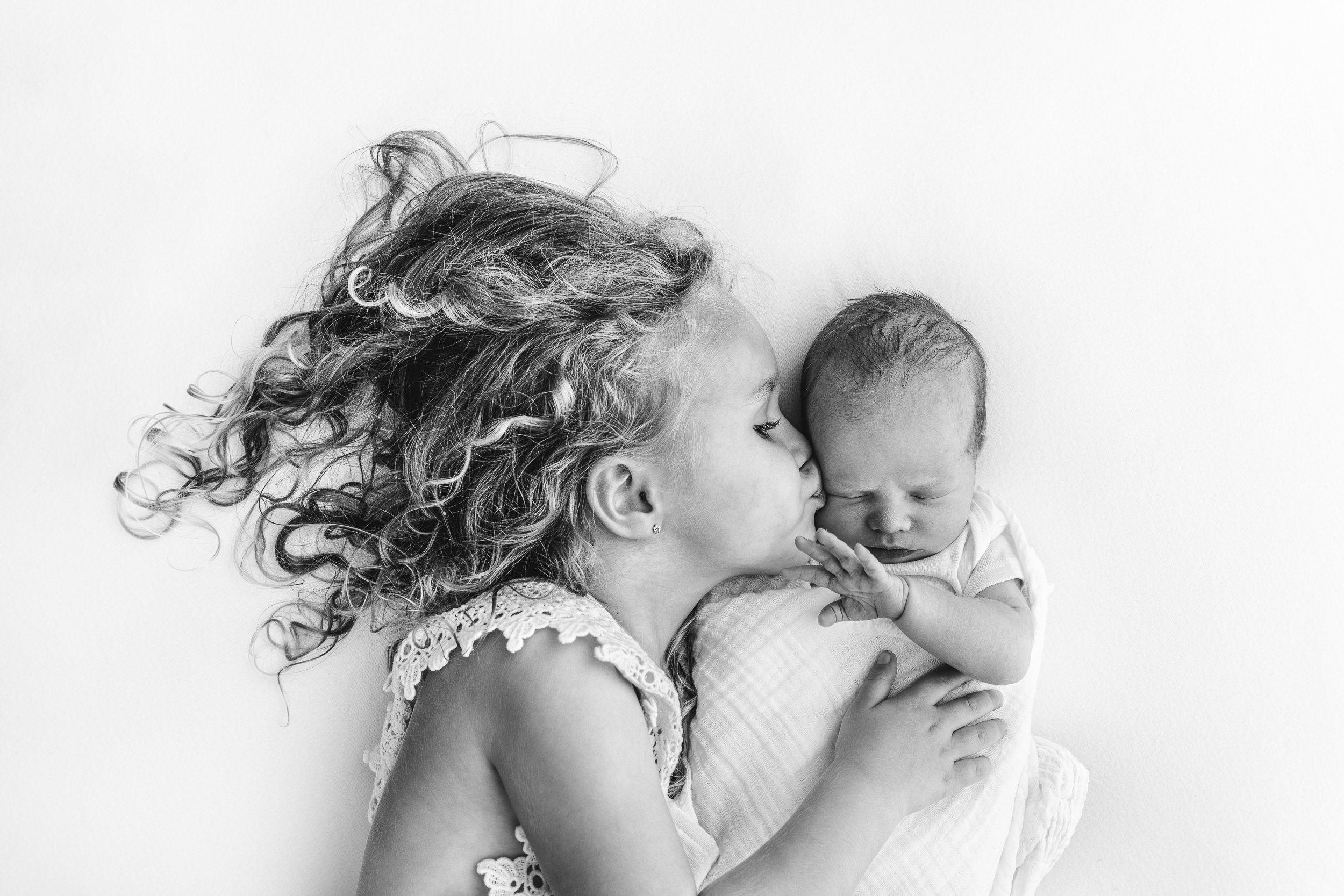  New Jersey photographer Nicole Hawkins Photography captures a big sister kissing her baby sister during a stunning studio session with all white. kissing baby sister older sisters kissing baby curly hair girl black and white pic #nicolehawkinsphotog