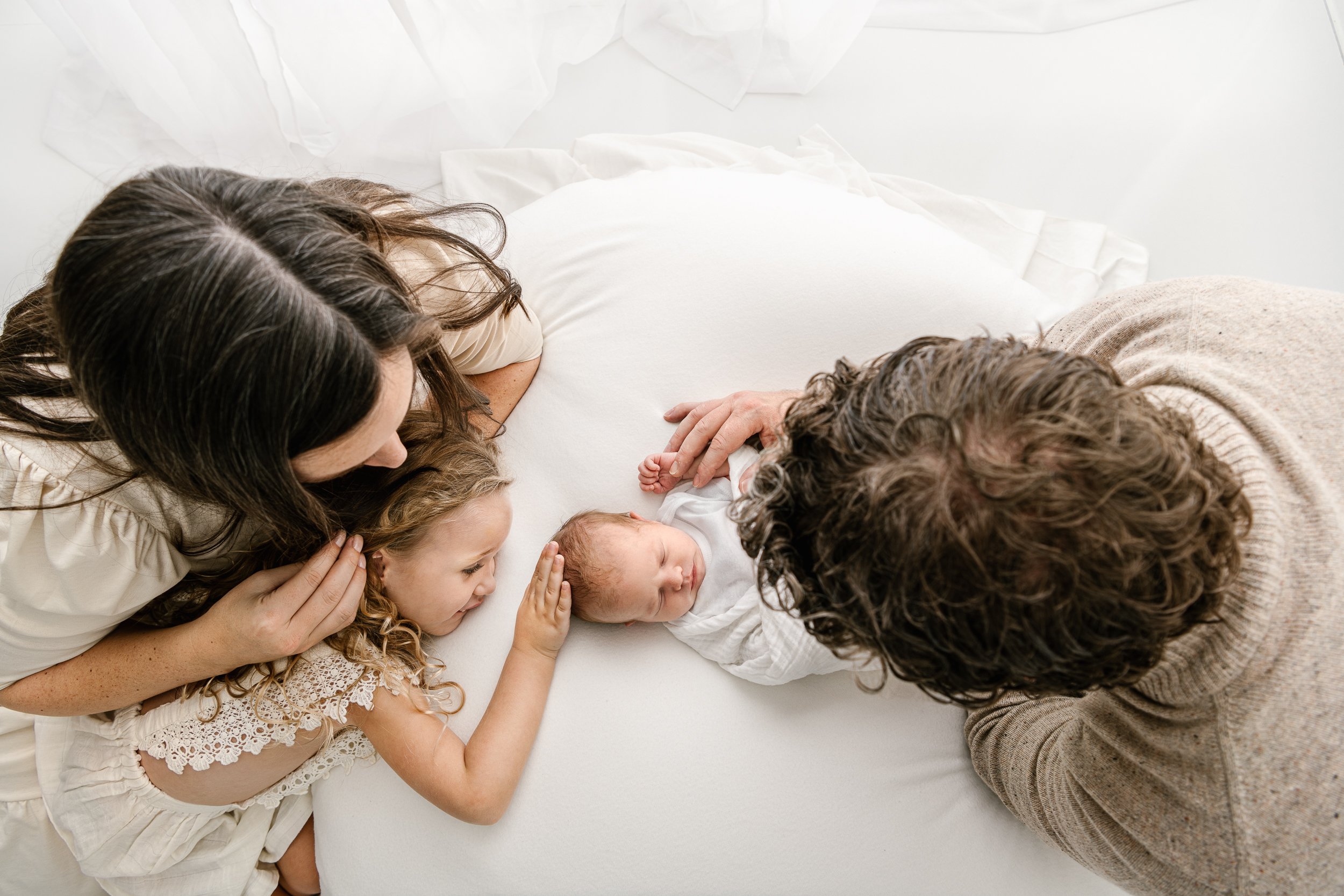  A beautiful aerial view of a family of four all doting on the sweet newborn baby girl on white bedding in a studio in New Jersey by Nicole Hawkins Photography. family of four stunning family of four studio portrait family studio poses #nicolehawkins