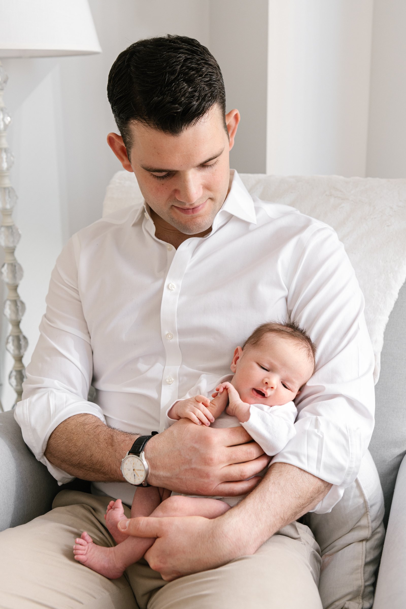  Father in a white button-up shirt and dark brown hair looks down at his new baby girl in his arms captured by Nicole Hawkins Photography in New Jersey. baby and father men photography style for in-home session baby girl #nicolehawkinsphotography #ni